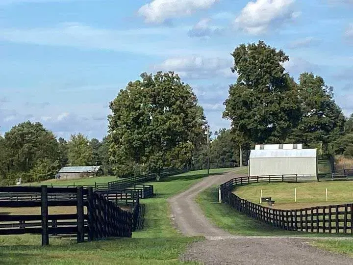 Curving dirt driveway between black fences in a green pasture under a partly cloudy sky.