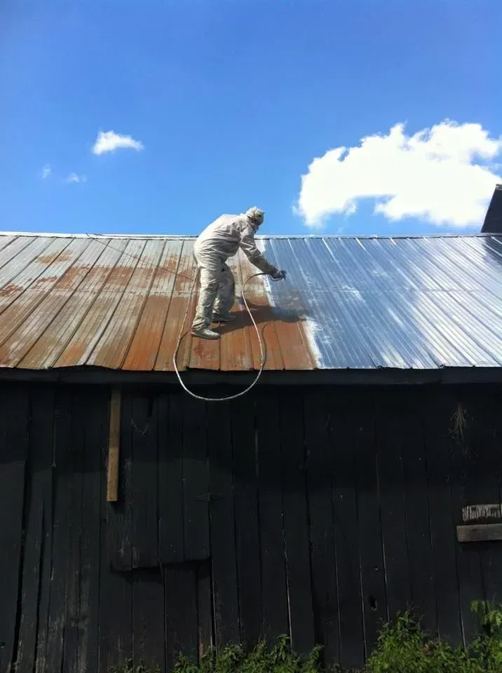 Person in protective suit spraying a rusty metal roof beside a dark building under a blue sky.