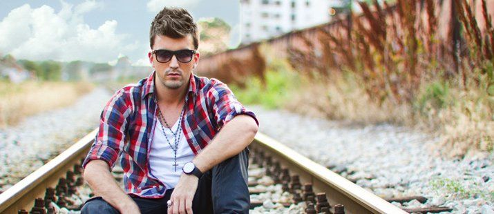 A man is sitting on train tracks wearing sunglasses and a plaid shirt.