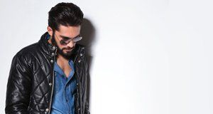 A man wearing a leather jacket and sunglasses is leaning against a white wall.