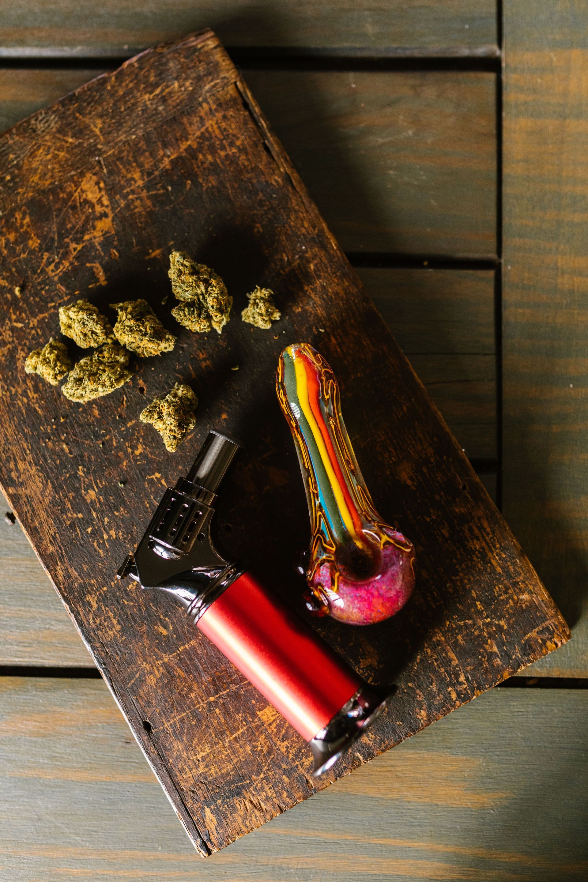 Red lighter and rainbow glass pipe on a dark tray with cannabis buds.