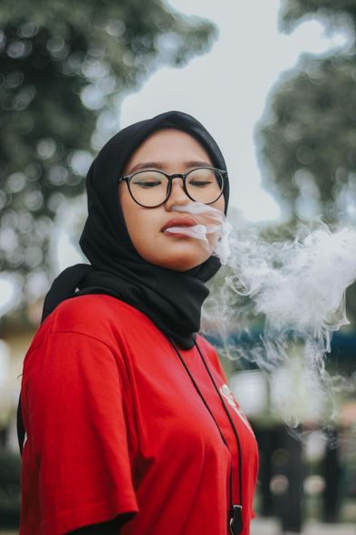 A woman wearing glasses and a hijab is blowing smoke out of her mouth.