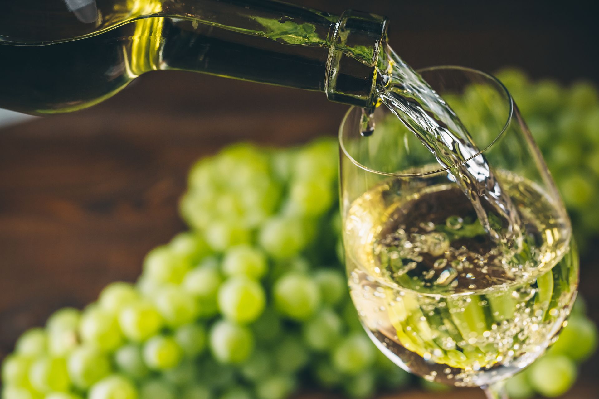 Wine being poured into a glass beside green grapes on a rustic table