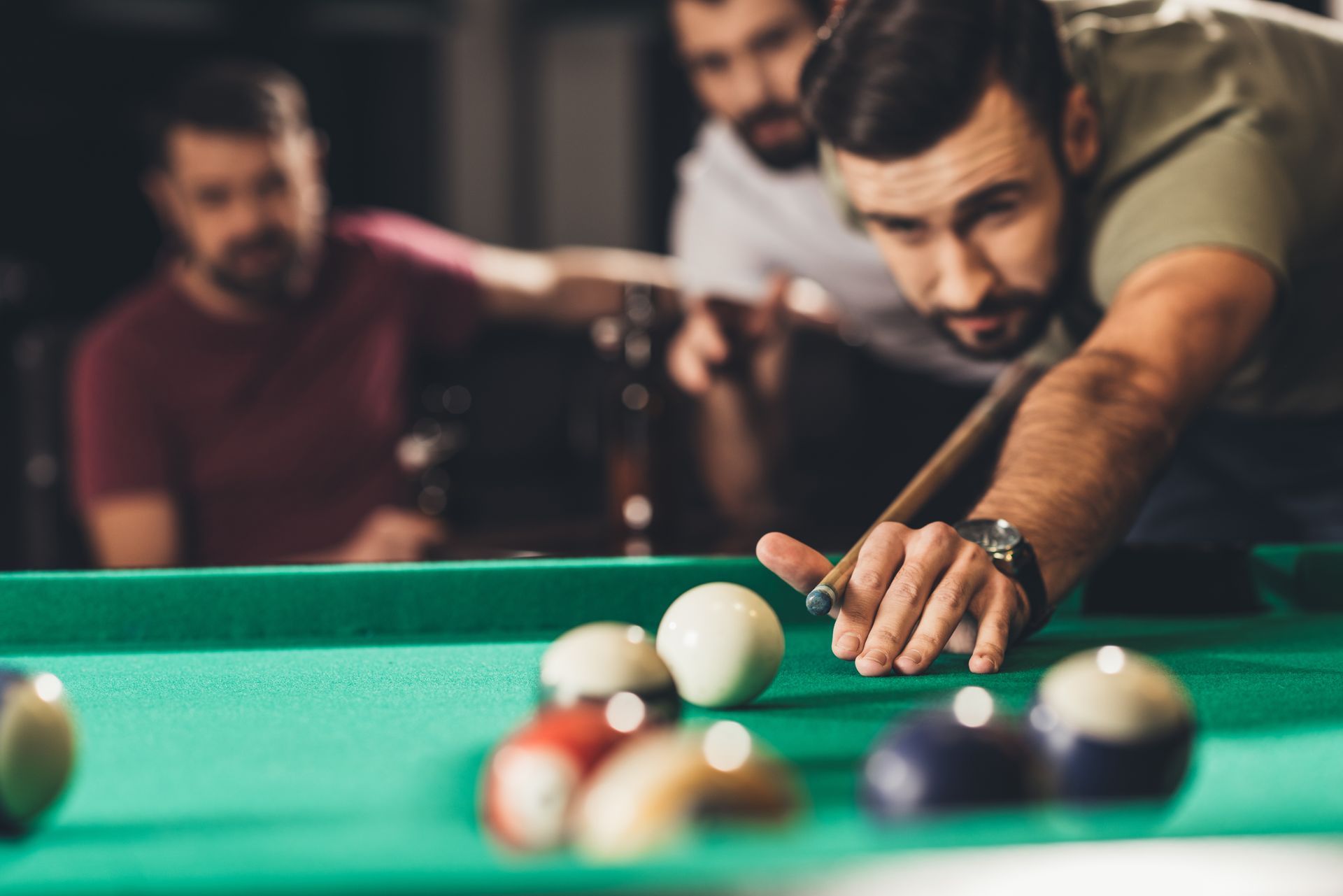 Man lining up a shot at a green pool table while two others watch in the background