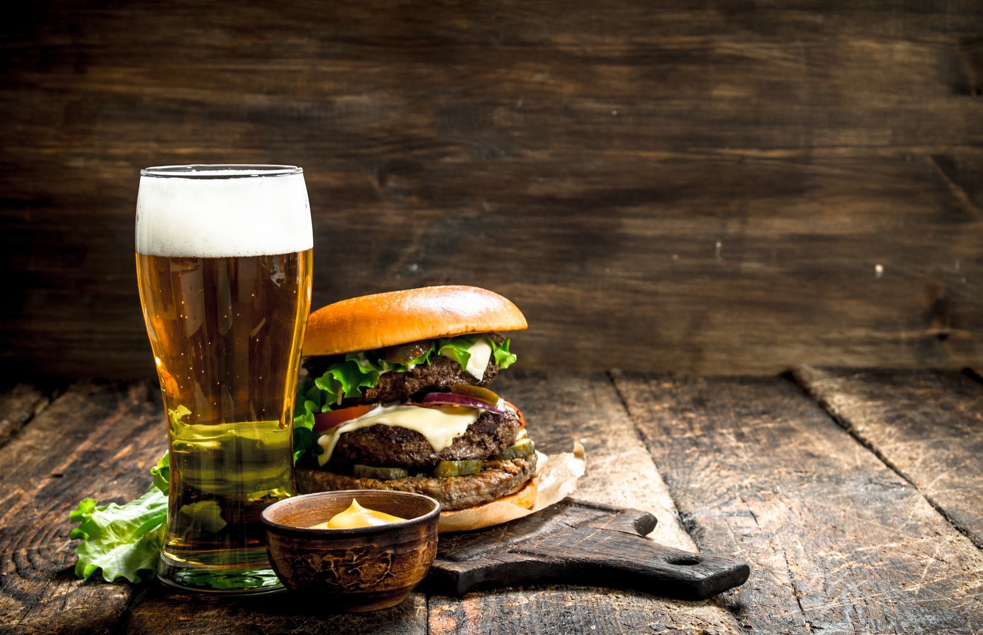 Beer and burger on a rustic wooden table, with a small bowl of sauce and leafy greens.
