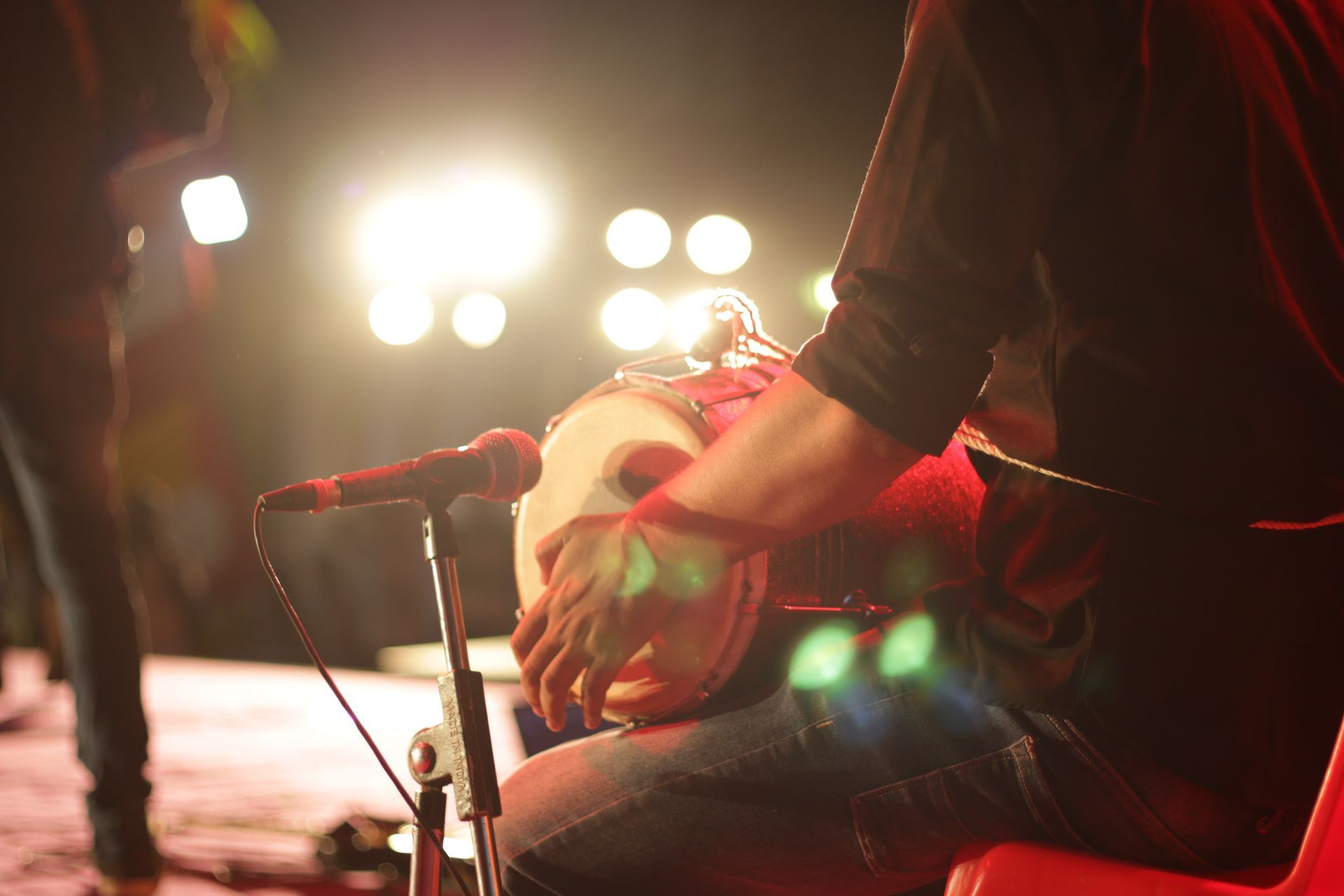 Musician seated on stage holding a microphone, with bright concert lights glowing behind them.