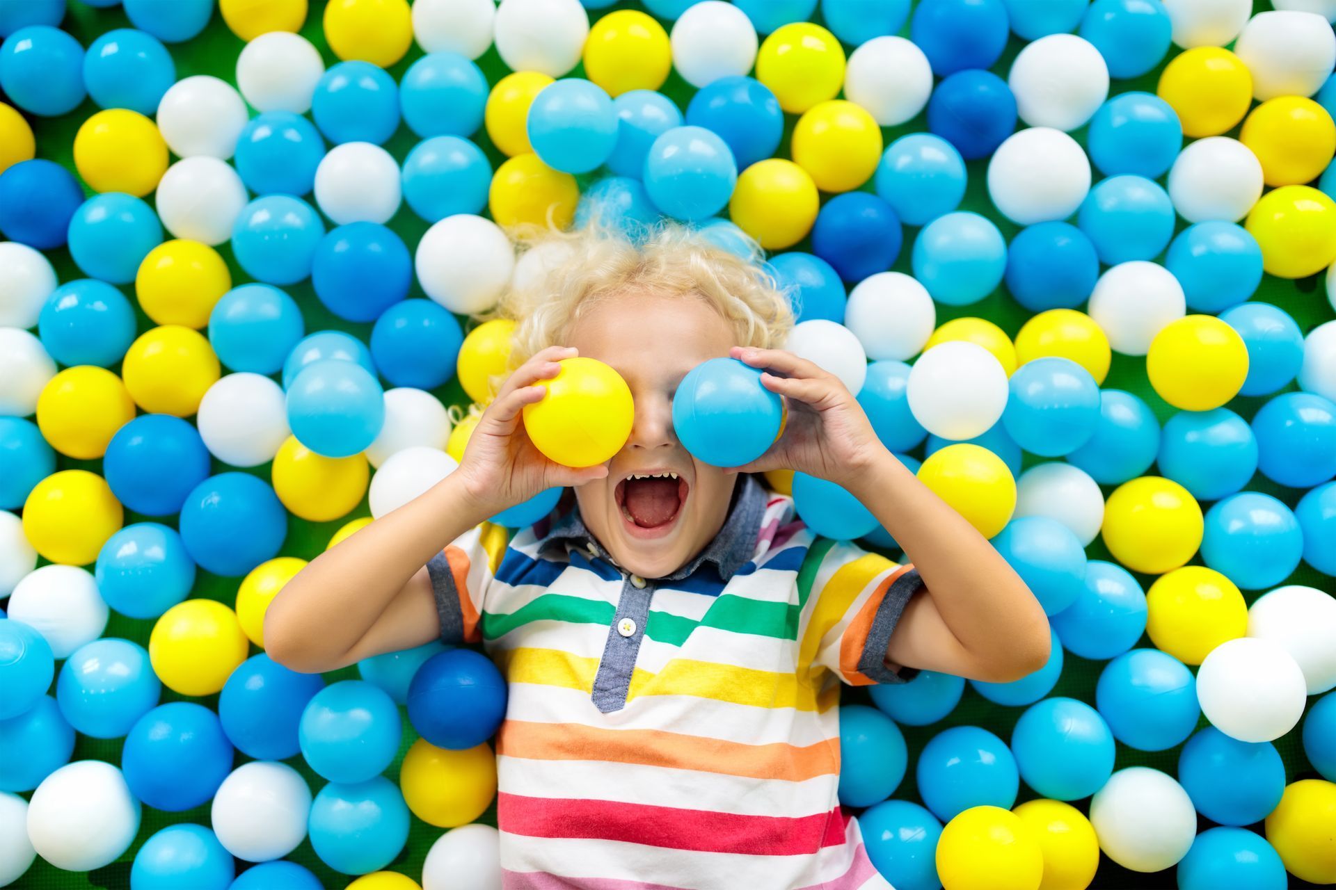 Person holding yellow and blue balls over eyes in a colorful ball pit, smiling
