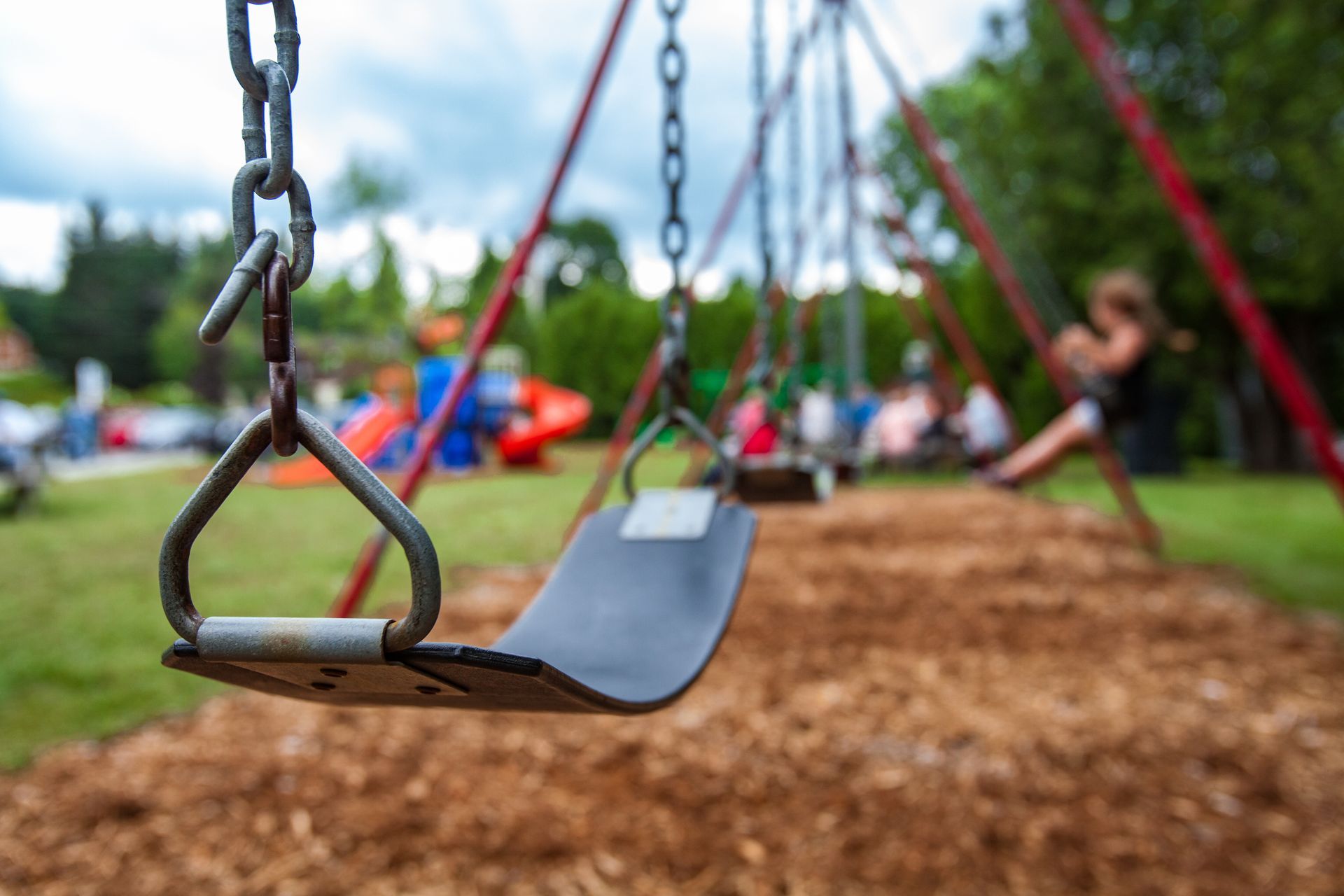 Close-up of a playground swing seat over wood chips, with children playing blurred in the background.