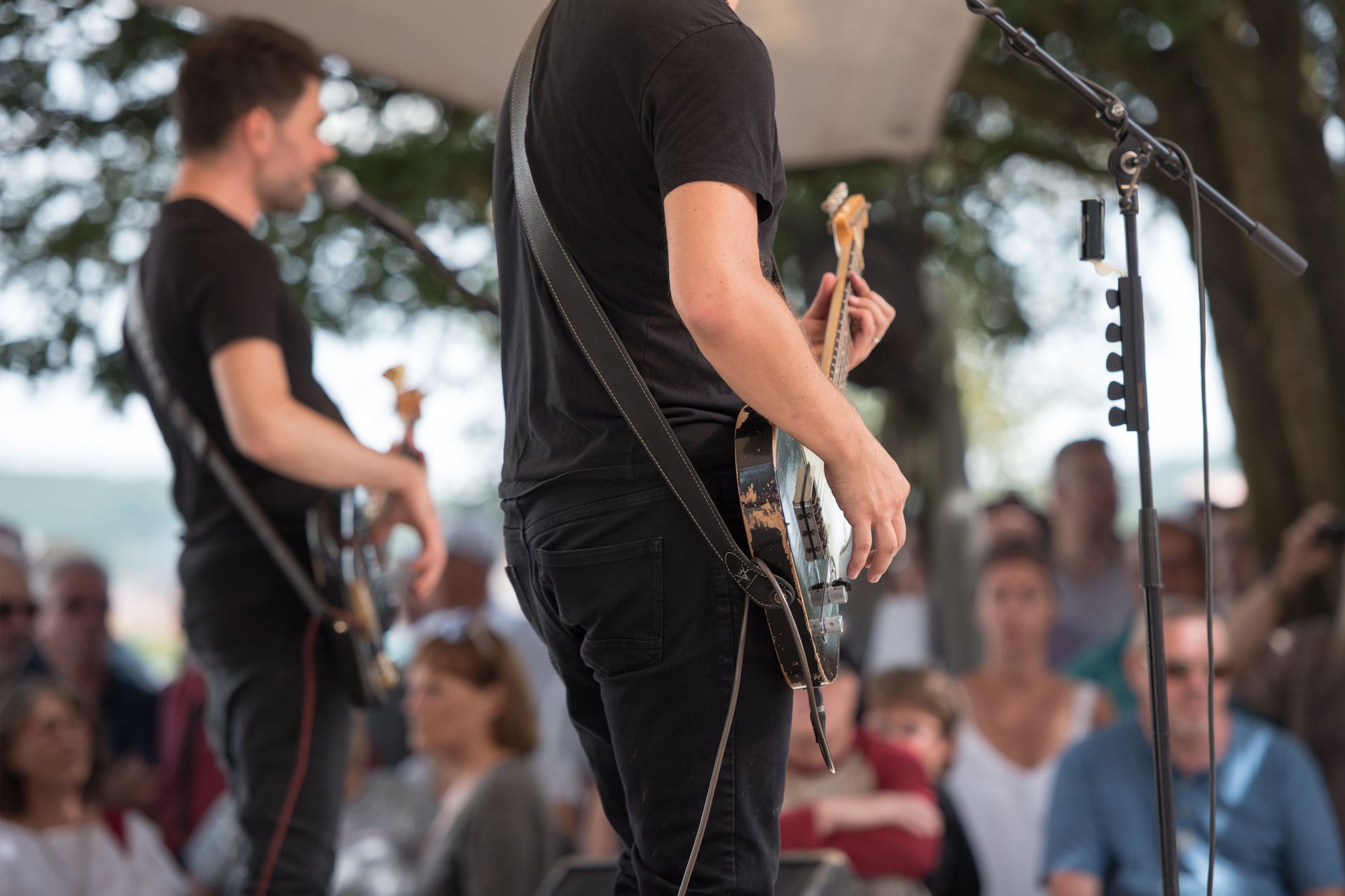 Musicians performing on an outdoor stage, with a guitarist in the foreground and audience blurred behind.