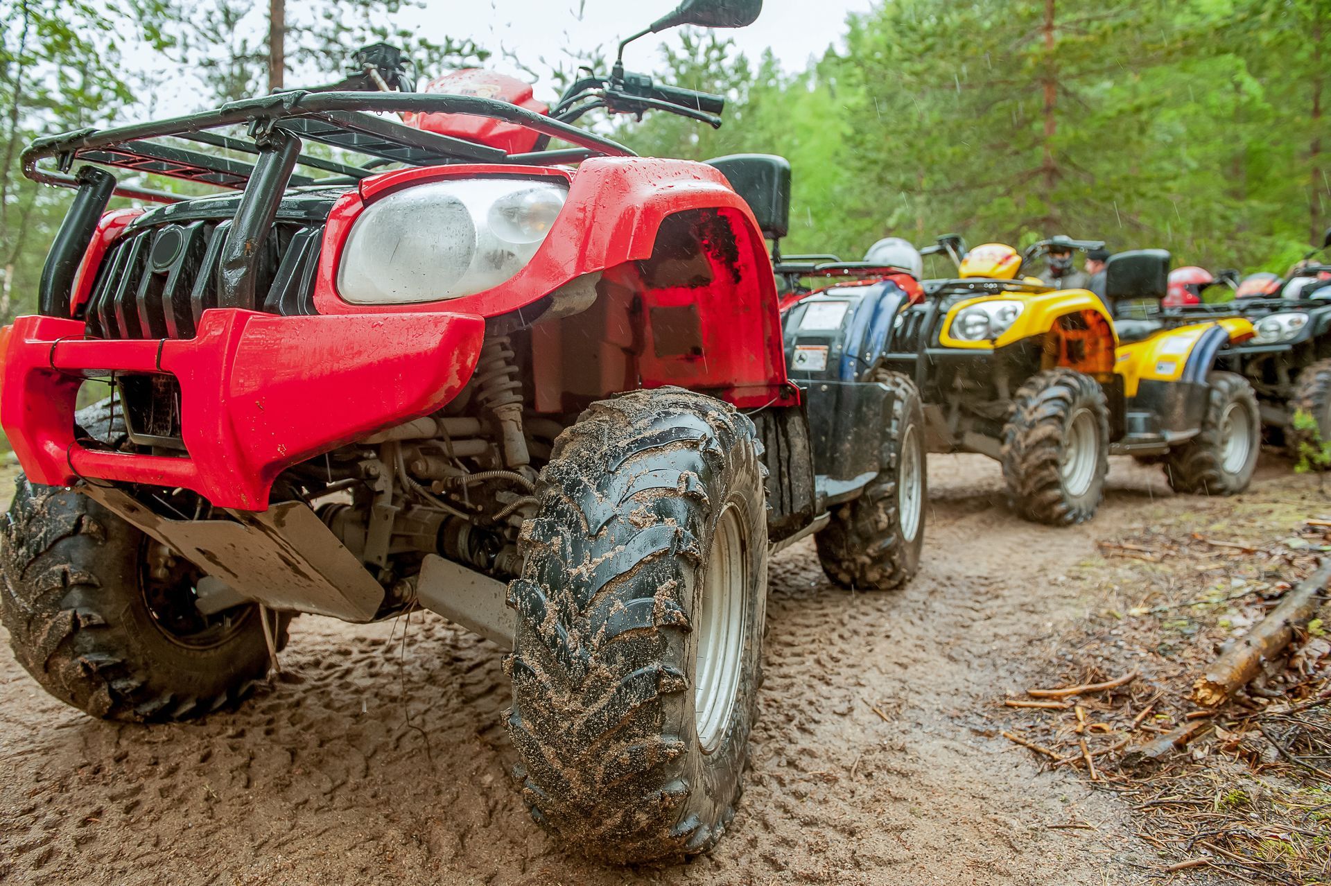 Red and yellow ATVs lined up on a muddy forest trail, with the front vehicle closest to the camera