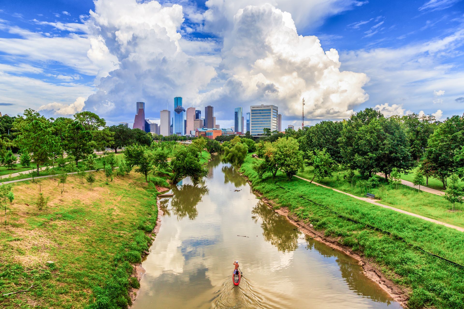 River winding through lush park toward a distant city skyline under large white clouds.