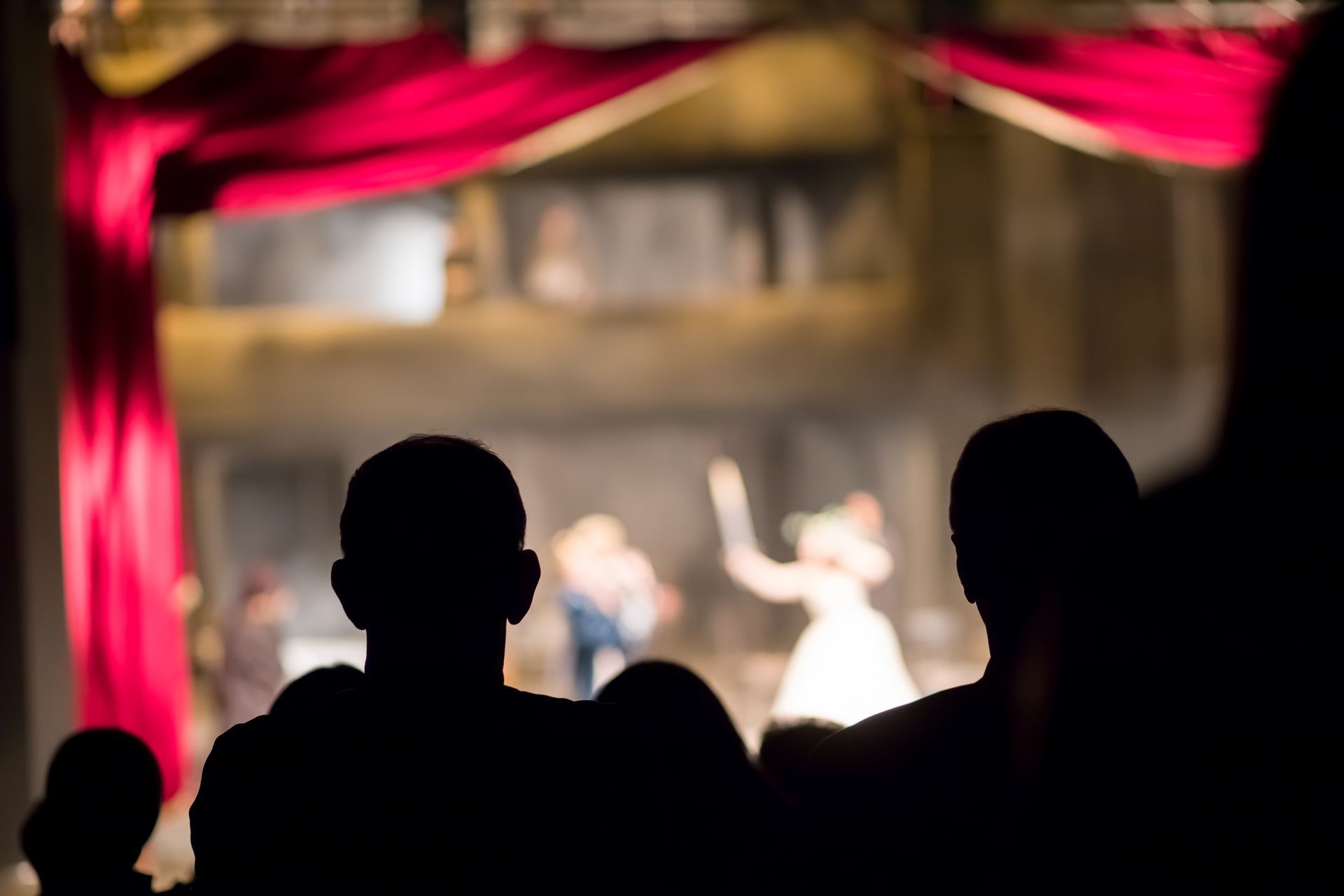 Audience silhouettes watching a brightly lit stage framed by red curtains