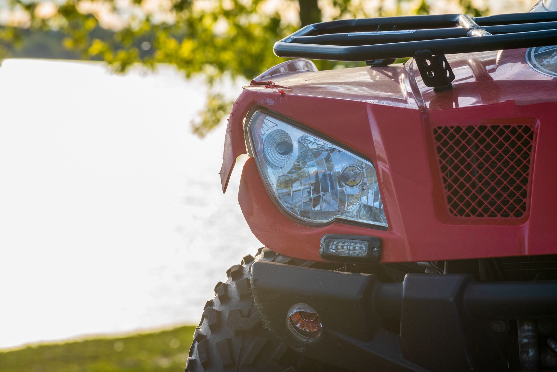 Close-up of a red ATV’s front headlight and grille, with a black rack and grassy outdoor background