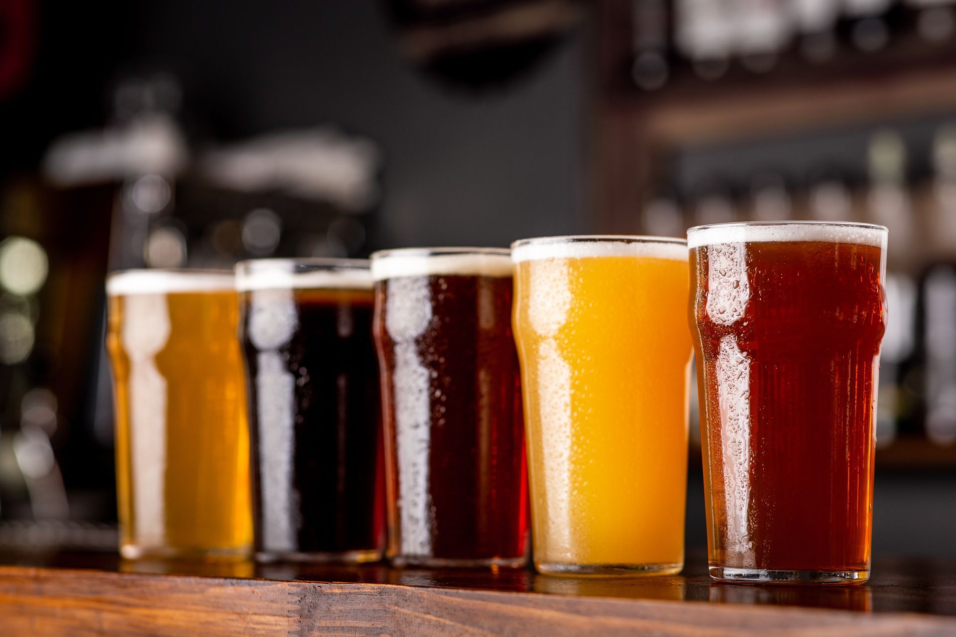 Five glasses of different beers lined up on a wooden bar counter, from light to dark amber.