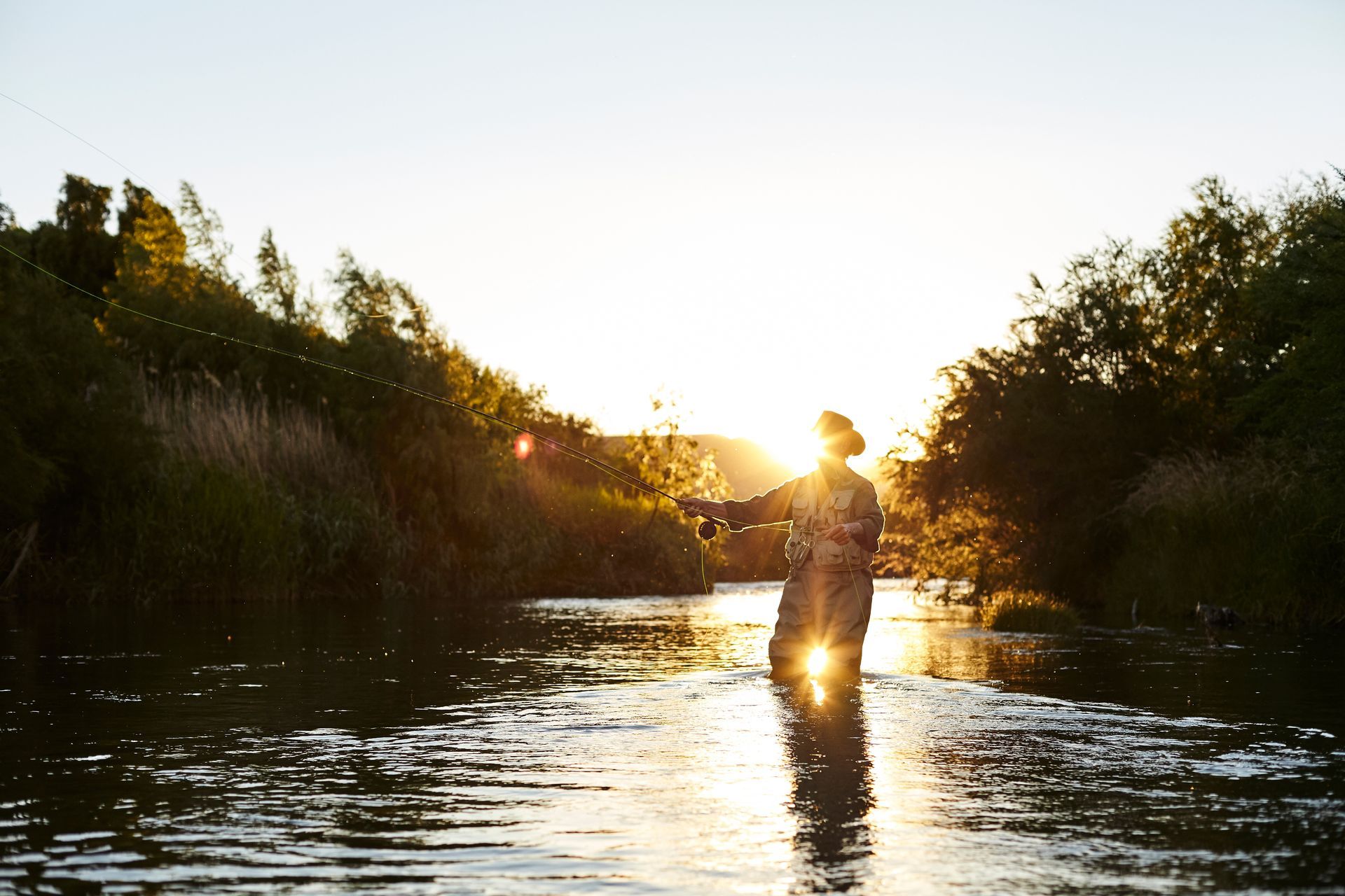 Person standing in a river at sunset, with warm light reflecting on the water and trees along the banks.