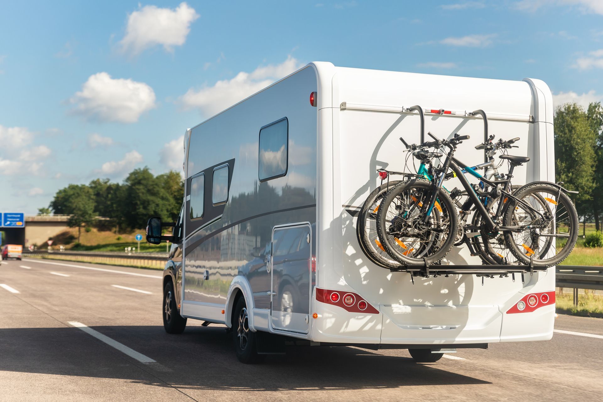 White RV towing bicycles on a rear rack, driving on a sunny highway.