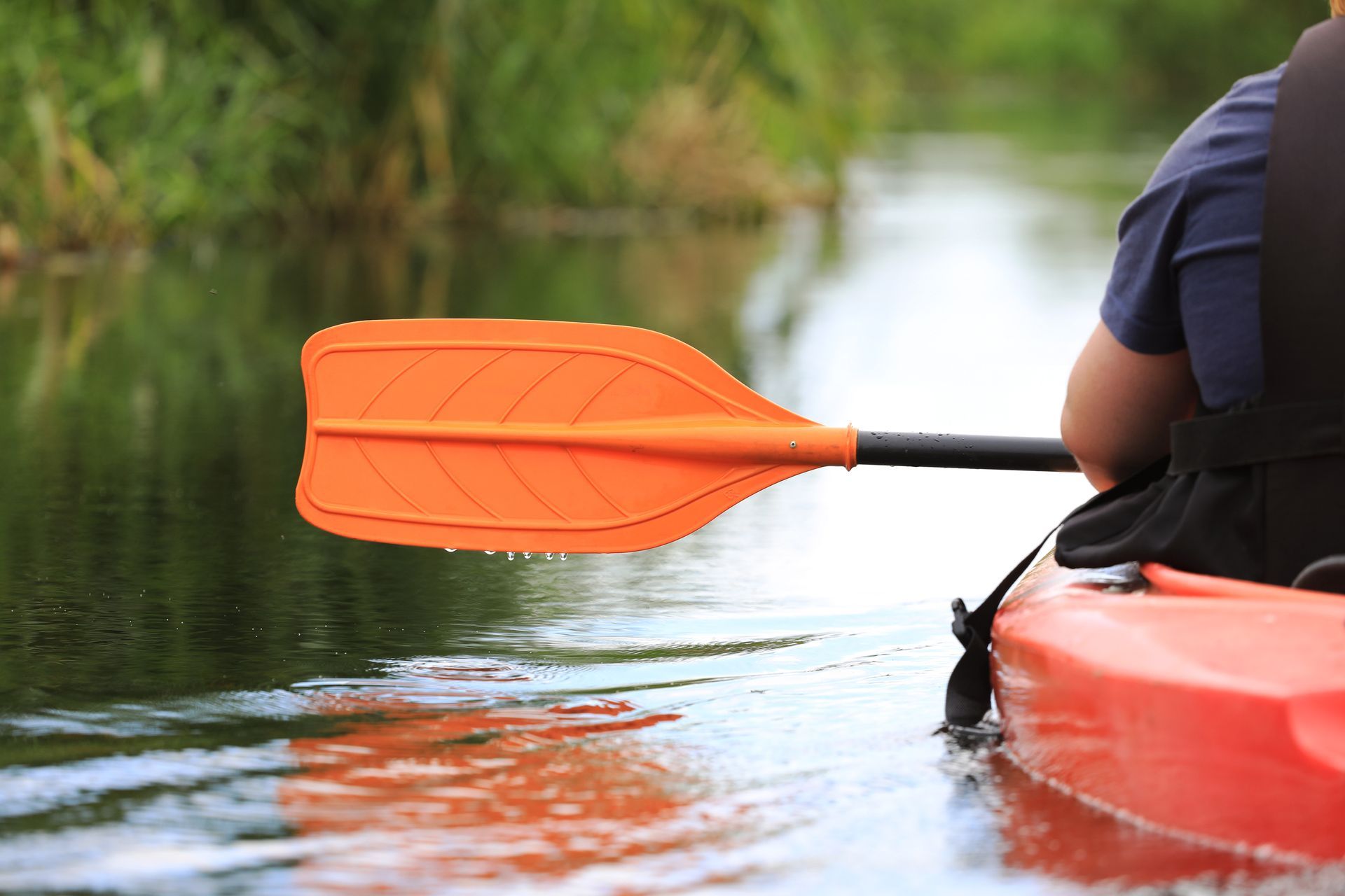 Orange kayak paddle on calm water beside a red kayak, with a blurred shoreline in the background