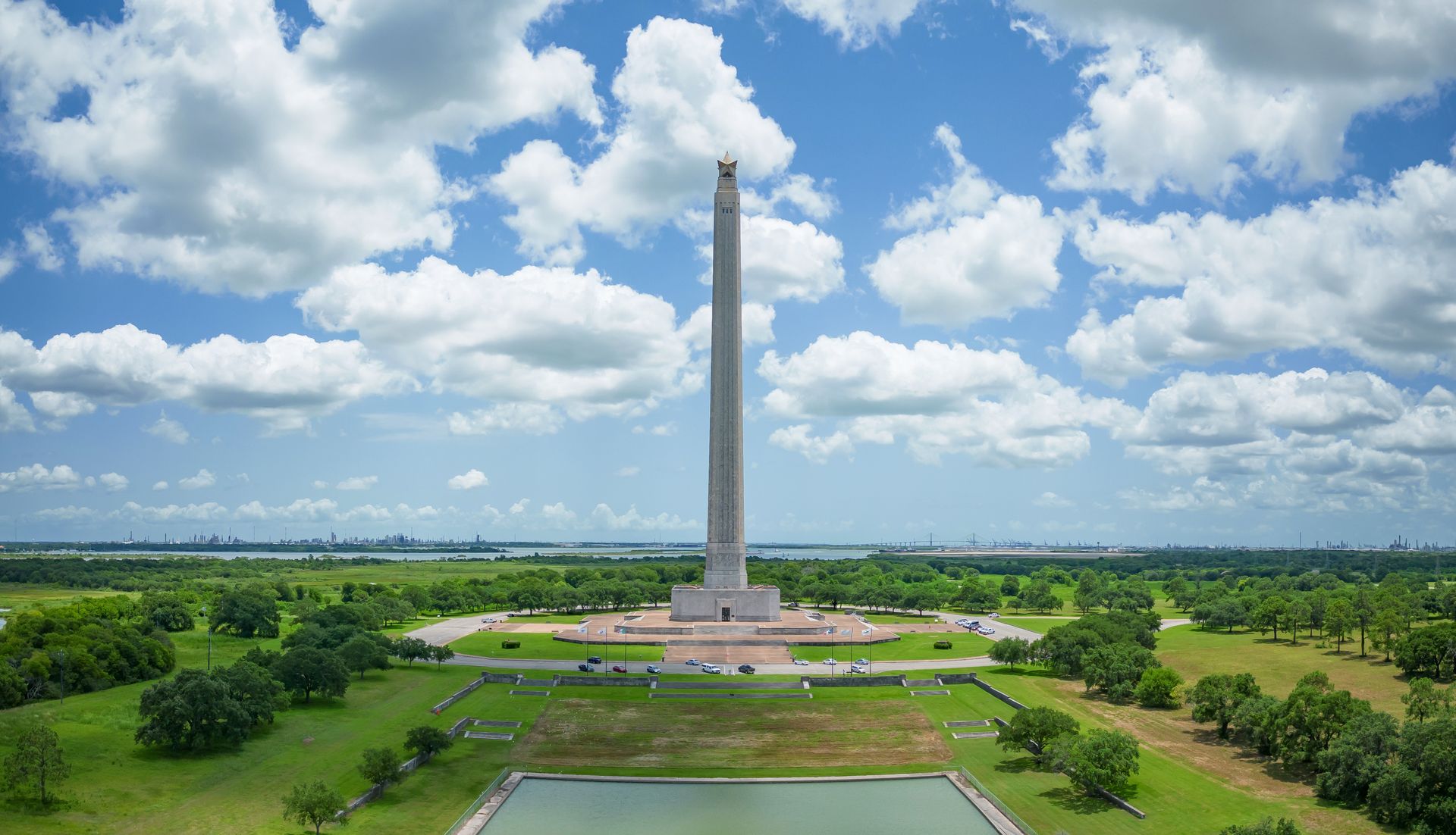 Tall white monument in a green park beneath a blue sky with clouds, viewed from a central path