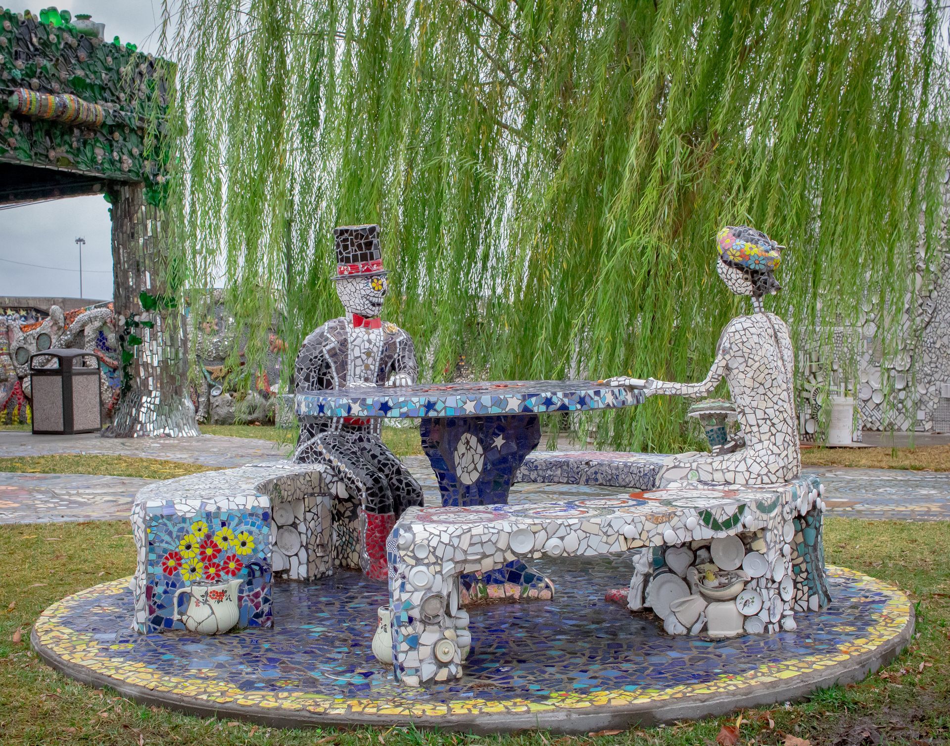 Two mosaic statues seated at a round table under a weeping willow in a garden