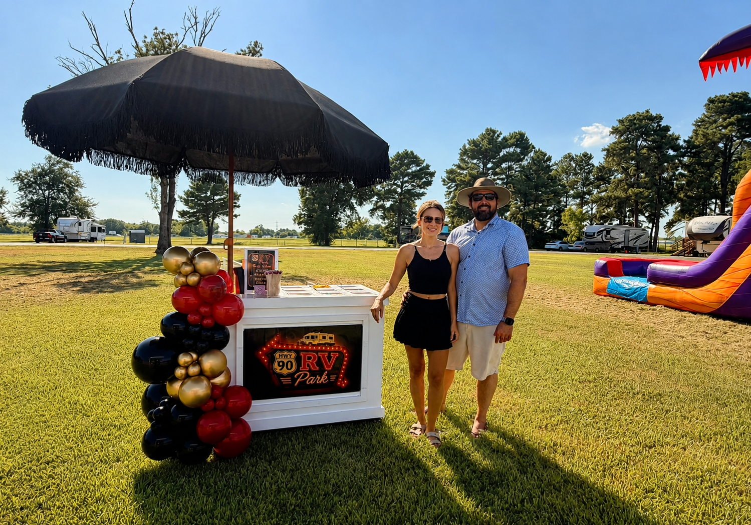 Two people standing beside a decorated outdoor party booth on a sunny lawn, with an inflatable slide in the background
