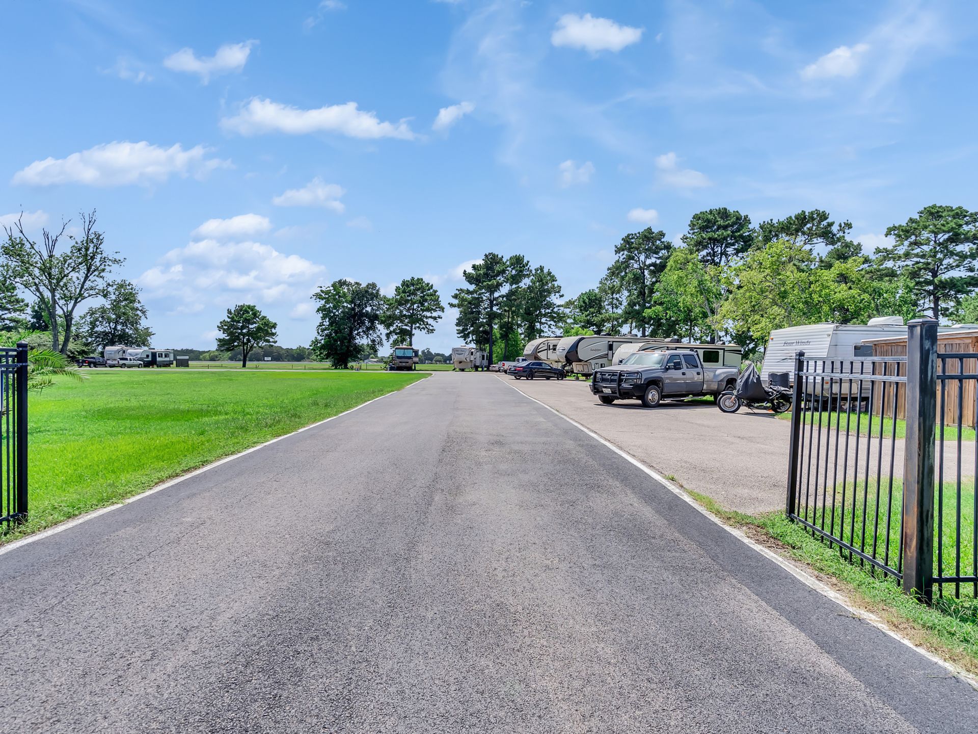 Wide paved road by green fields and parked cars under a sunny blue sky