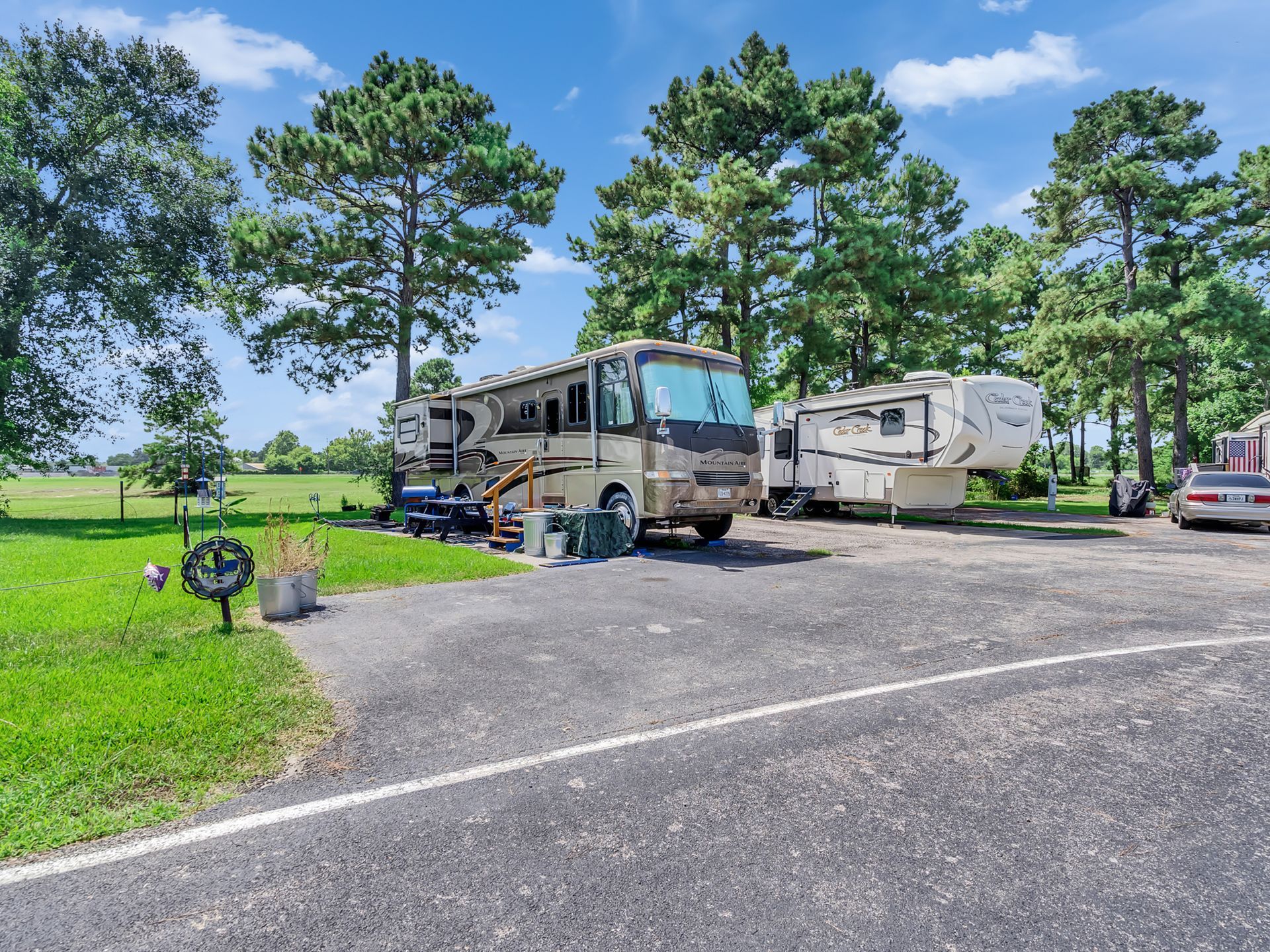 RVs parked at a wooded campground beside a paved road under a blue sky