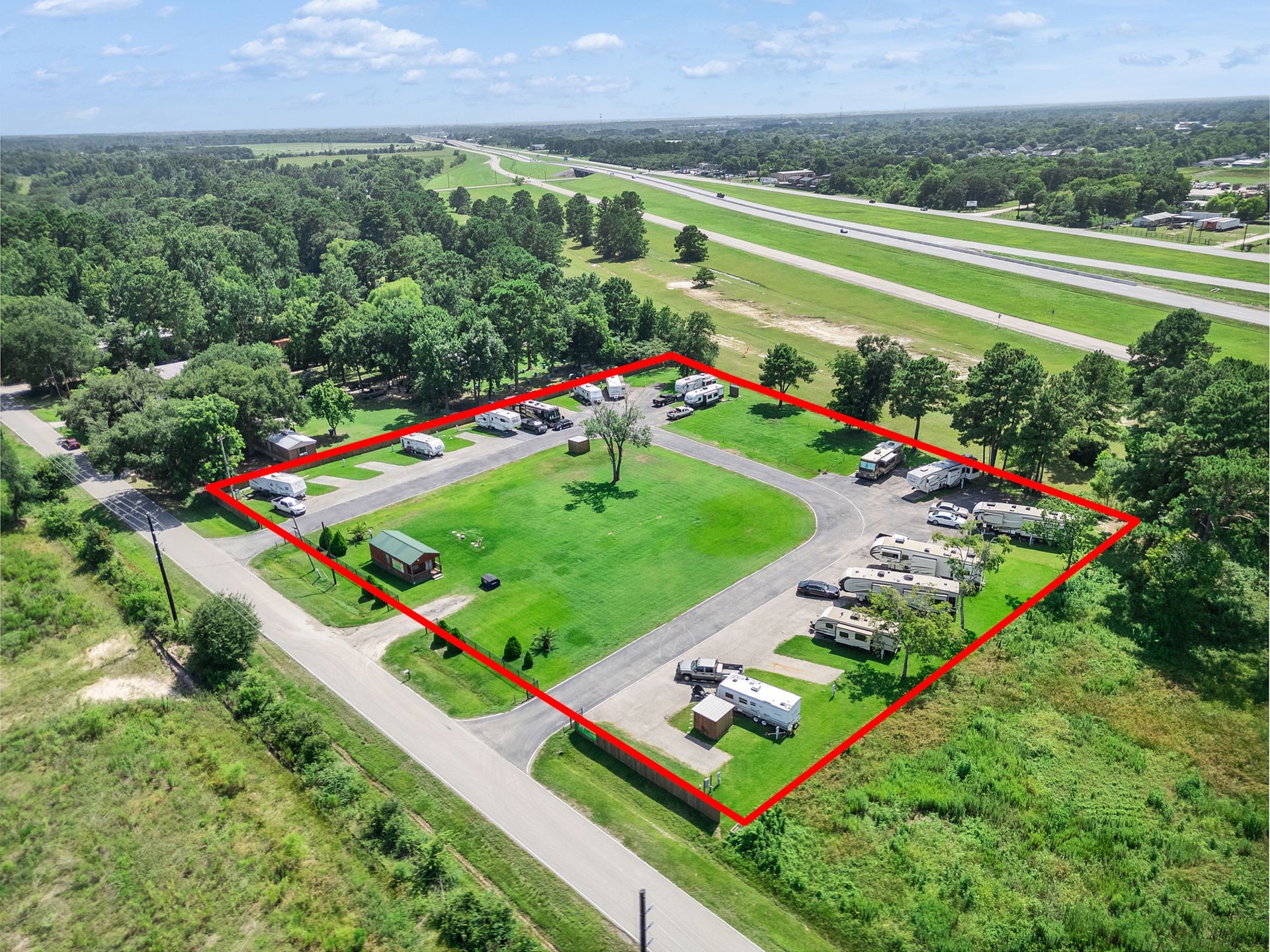 Aerial view of a fenced grassy park with a central tree, surrounded by roads and dense trees.