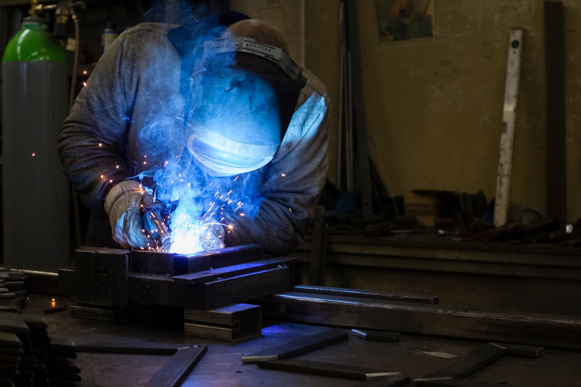 A welder in protective gear uses an electric arc to join metal pieces in a dim workshop with a green gas cylinder.