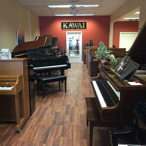 Piano showroom with several grand and upright pianos on a wood floor, Kawai sign on the back wall.