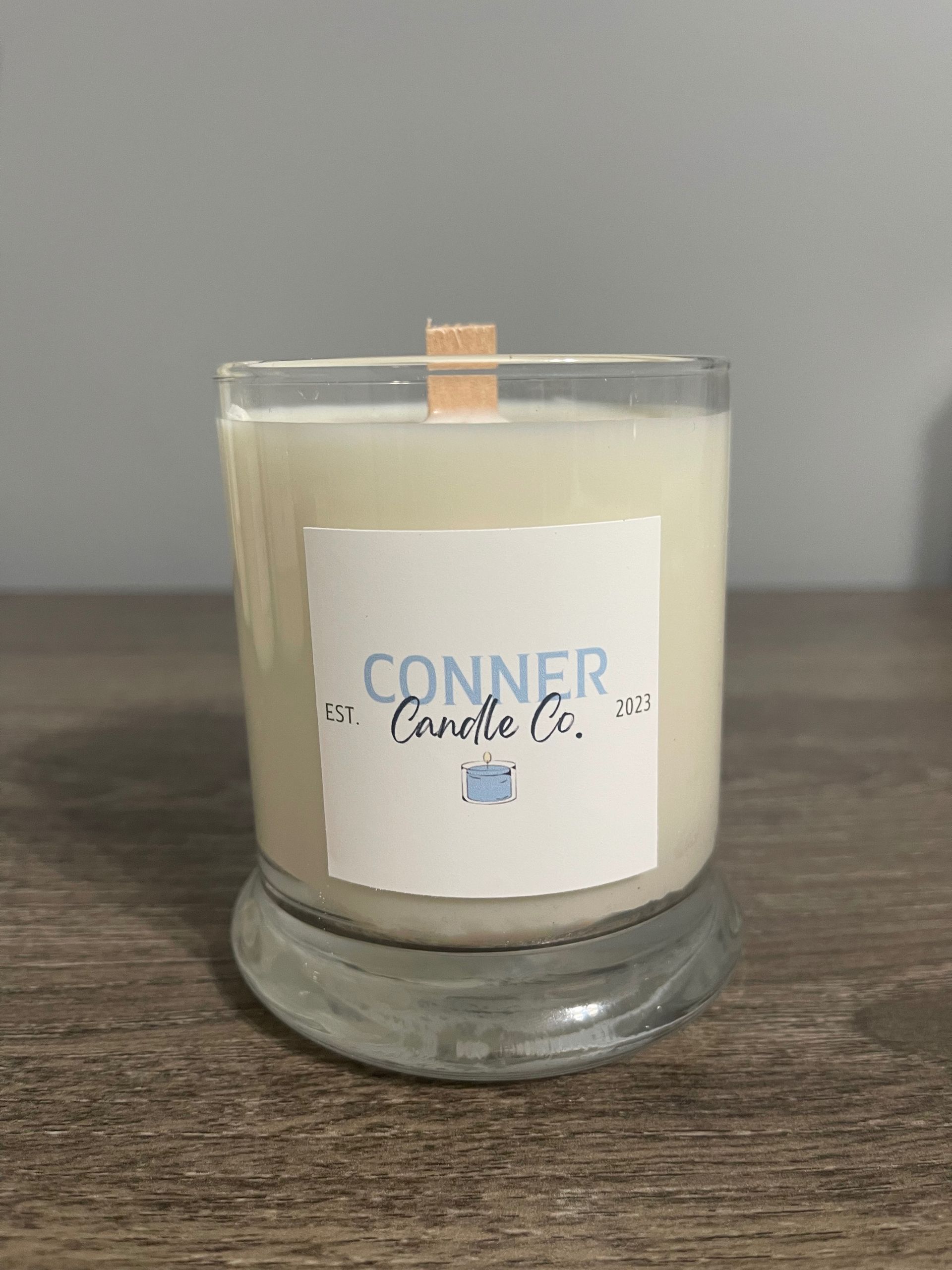 Candle in a clear glass jar with a wooden wick, labeled "Conner Candle Co." on a wooden surface.
