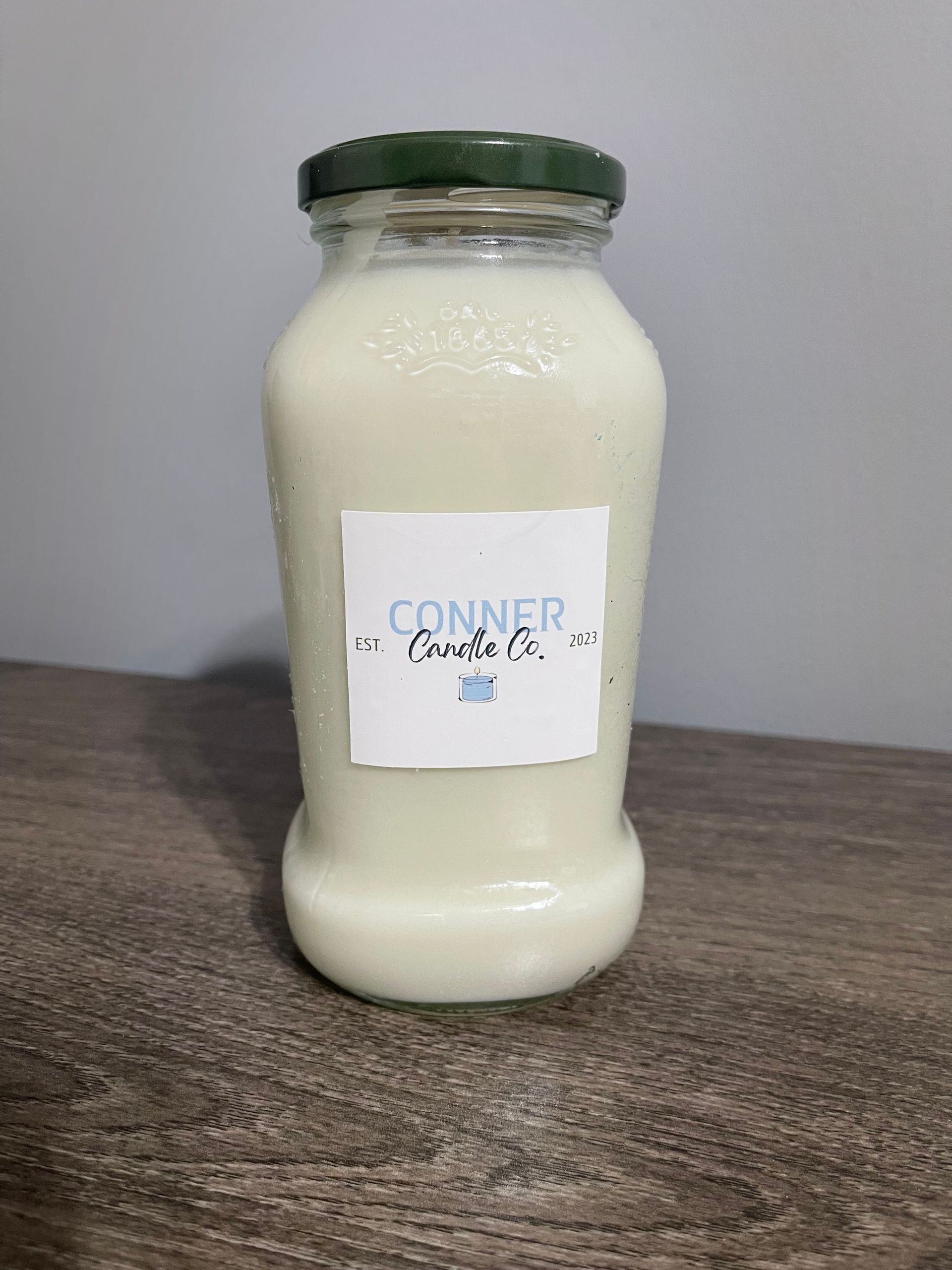 White candle in a glass jar with a green lid; label reads "Conner Candle Co." on a gray table.