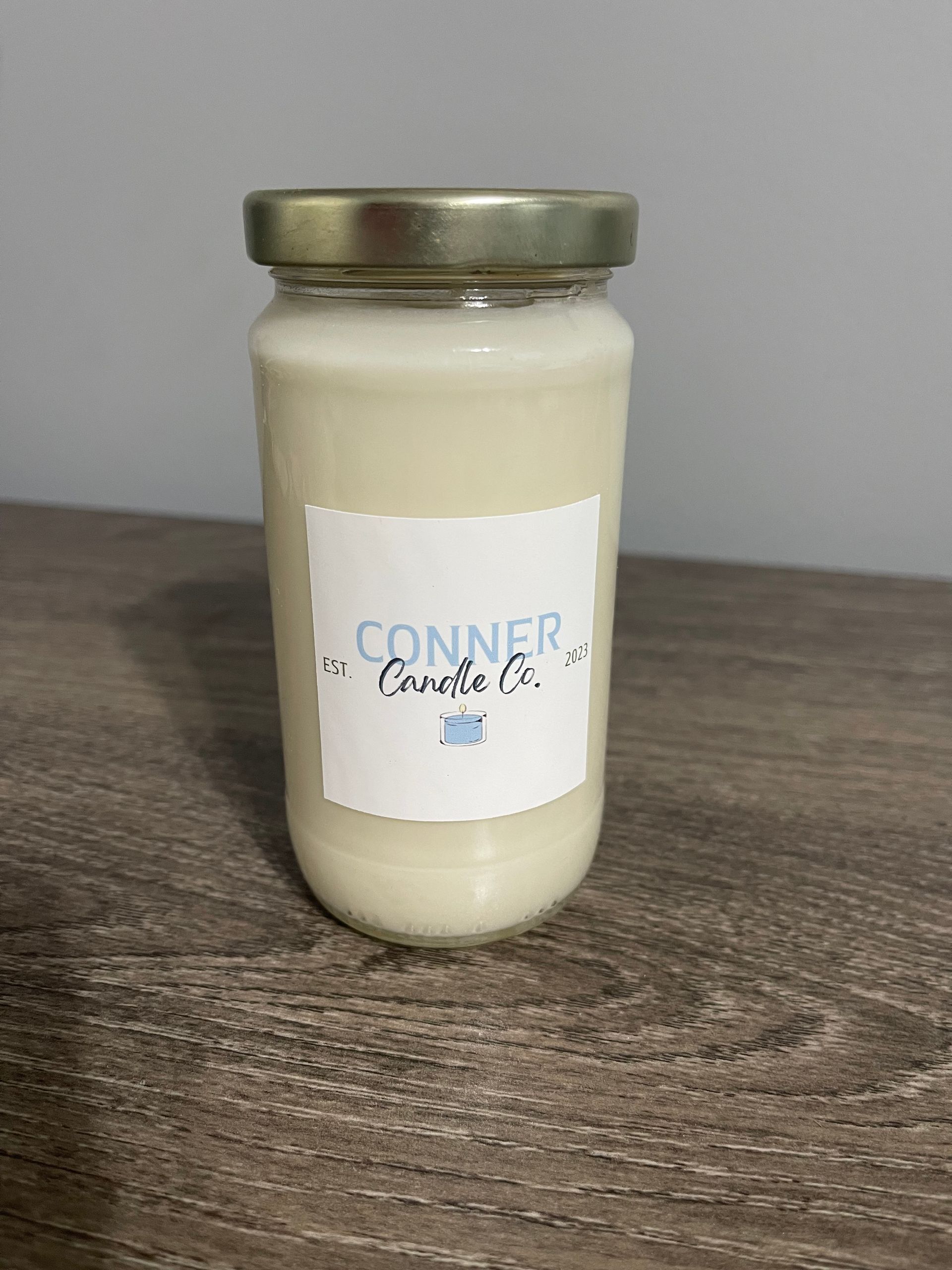 A white candle in a glass jar with a gold lid on a wooden surface. Label reads "Conner Candle Co."