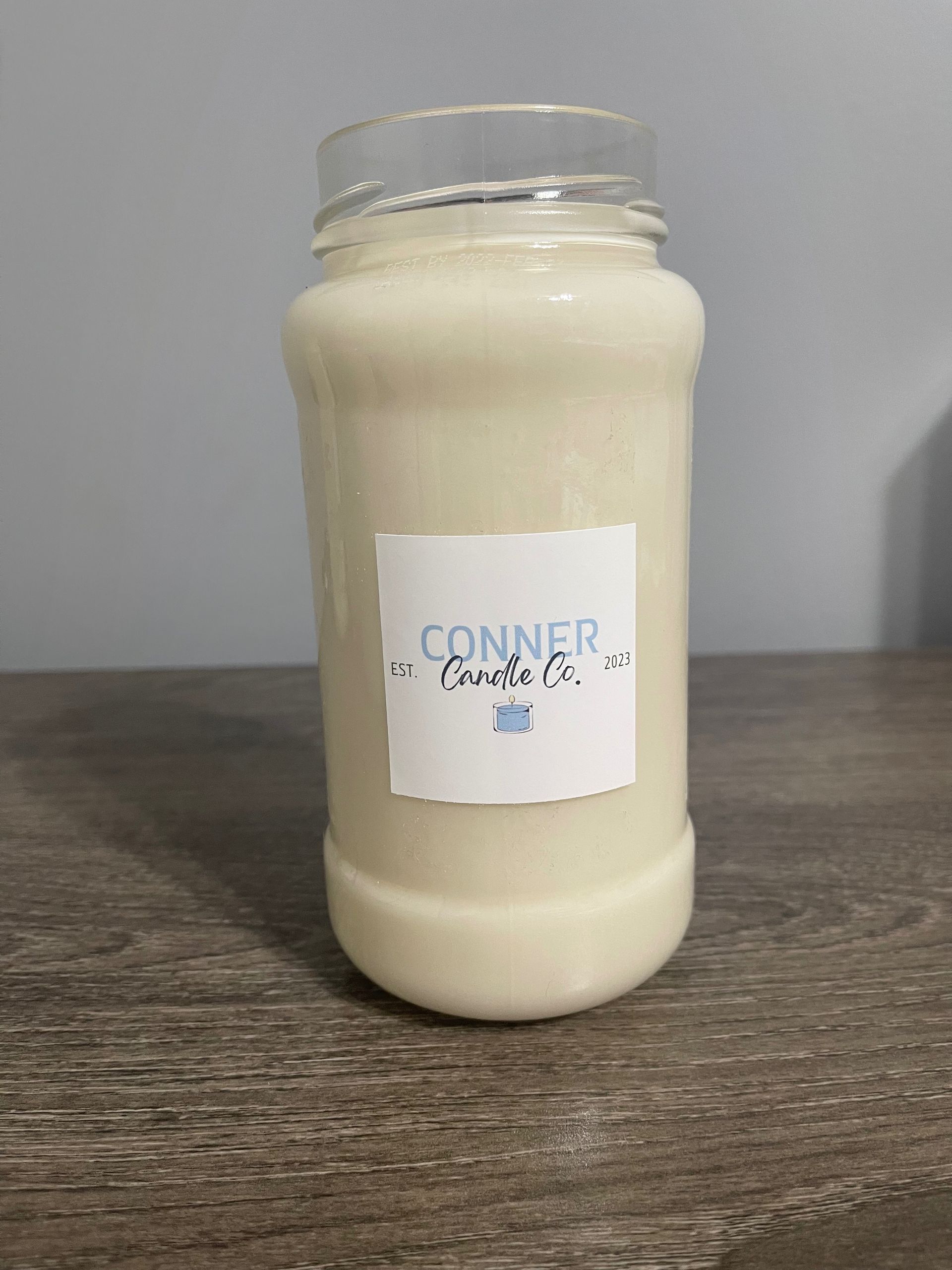 Jar of creamy liquid with a label that says "Conner Candles" on a wooden surface.