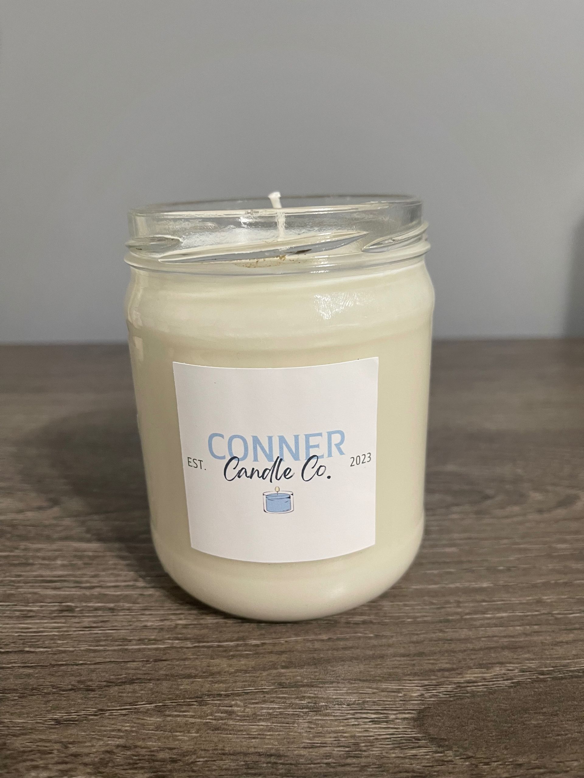 White candle in a mason jar with a label that says "Conner Candle Co." on a wooden surface.
