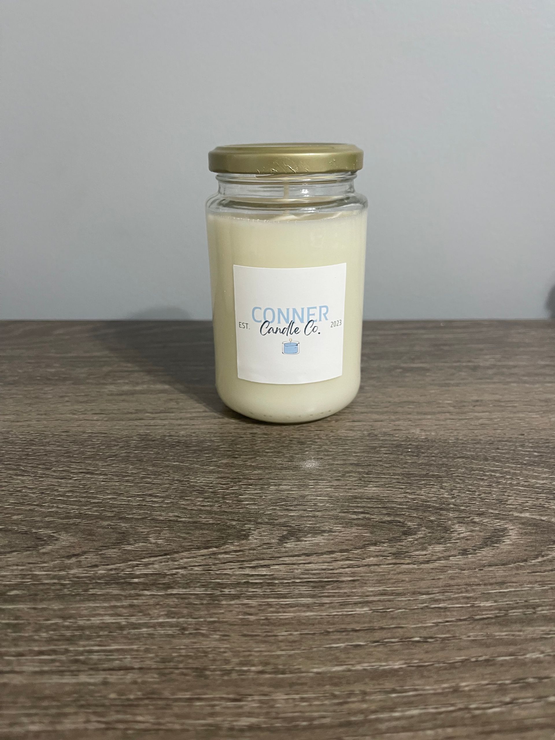 A candle in a glass jar, labeled "Conner" on a wooden surface with a neutral backdrop.