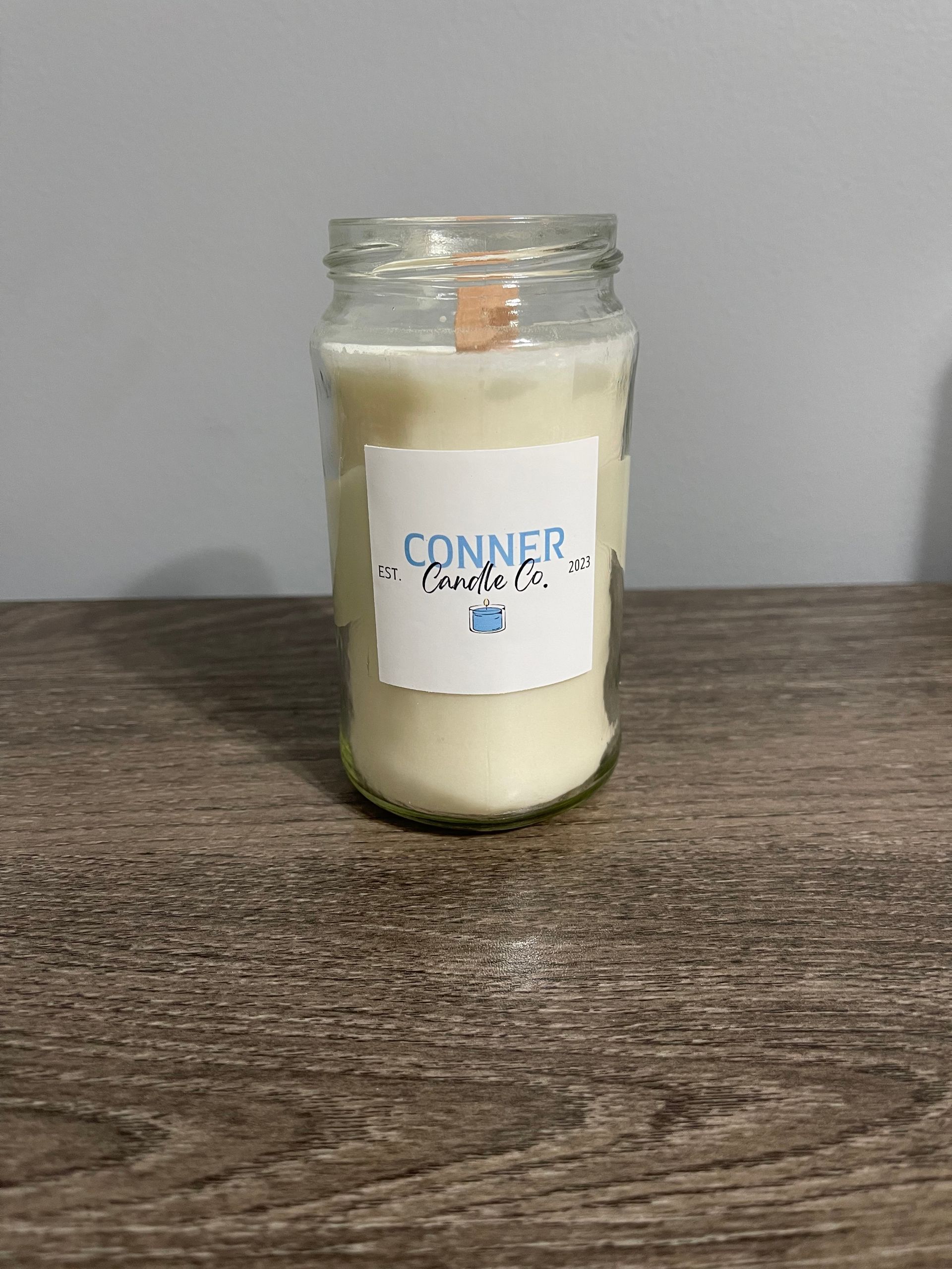 Glass jar candle with wooden wick and label "Conner Sucks Co." on wooden surface.