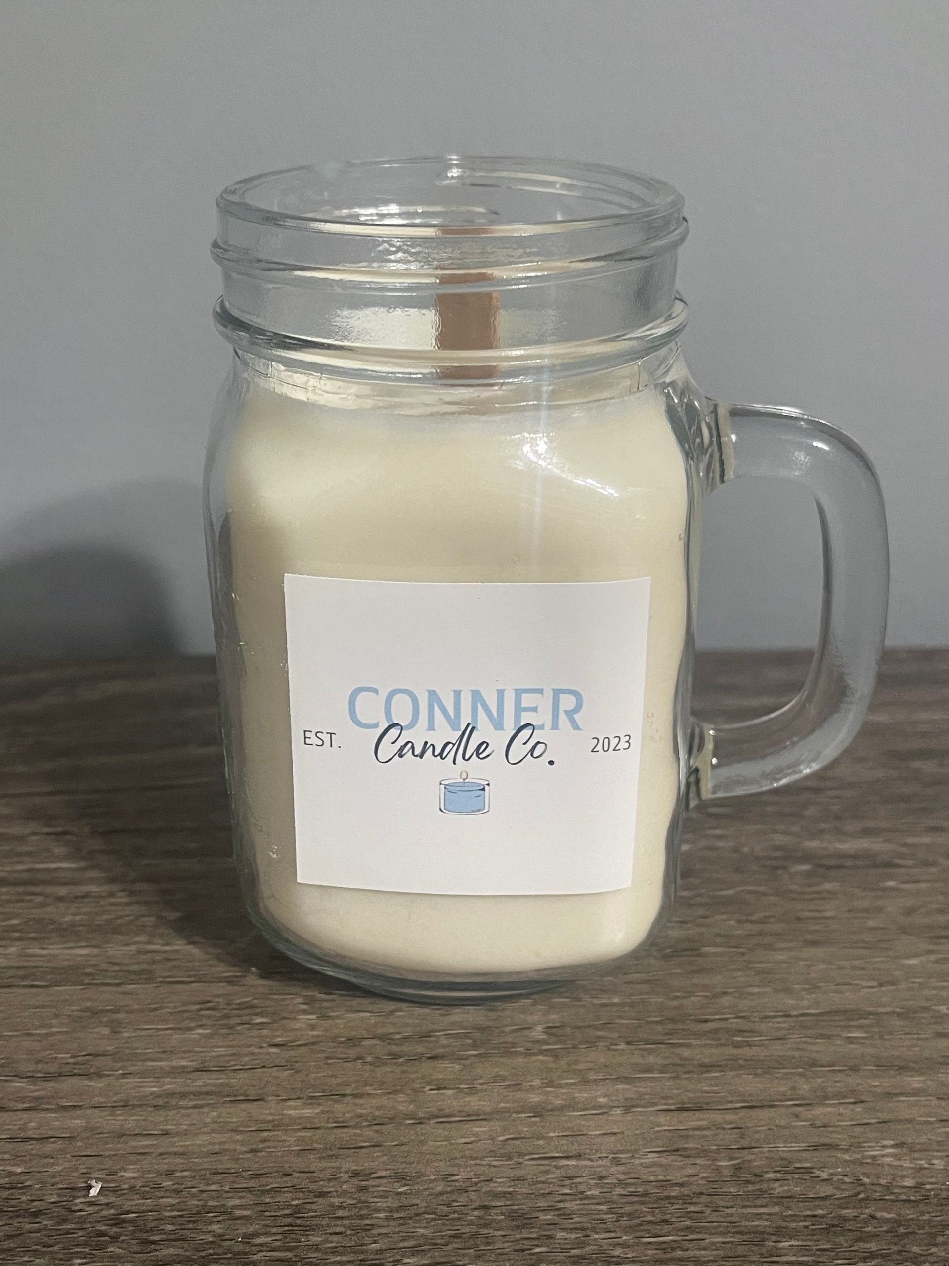 Cream-colored candle in a mason jar mug with wooden wick and label that reads "CONNER Candle Co." on a wooden surface.