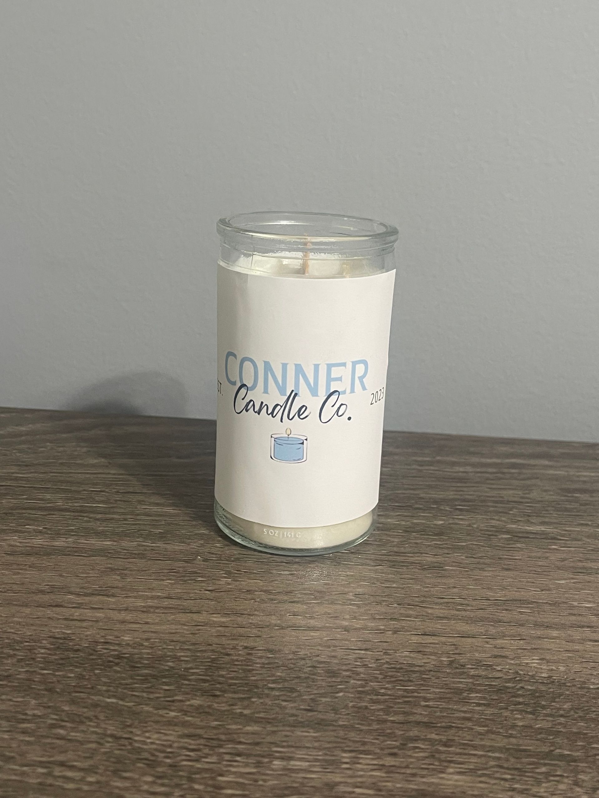 Candle in a clear glass jar with a white label that reads "CONNER Candle Co." on a wooden surface.