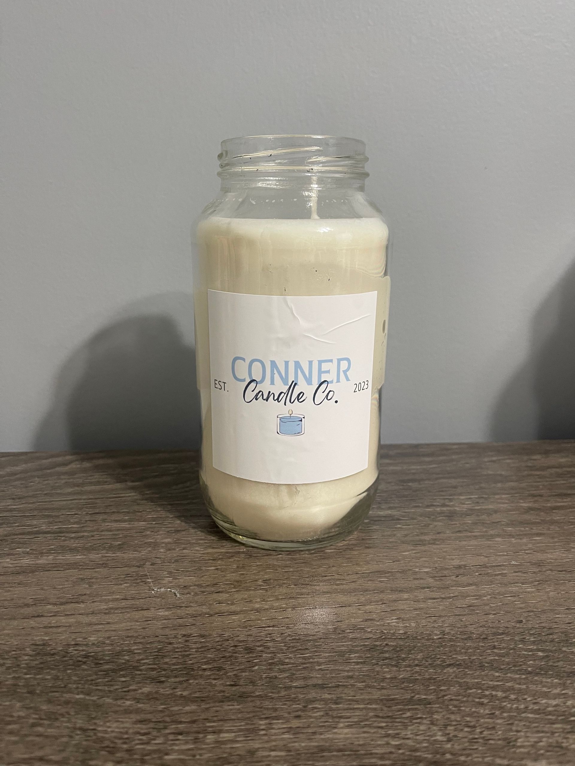 White candle in a glass jar with label that reads "CONNER Candle Co." on a wooden surface.
