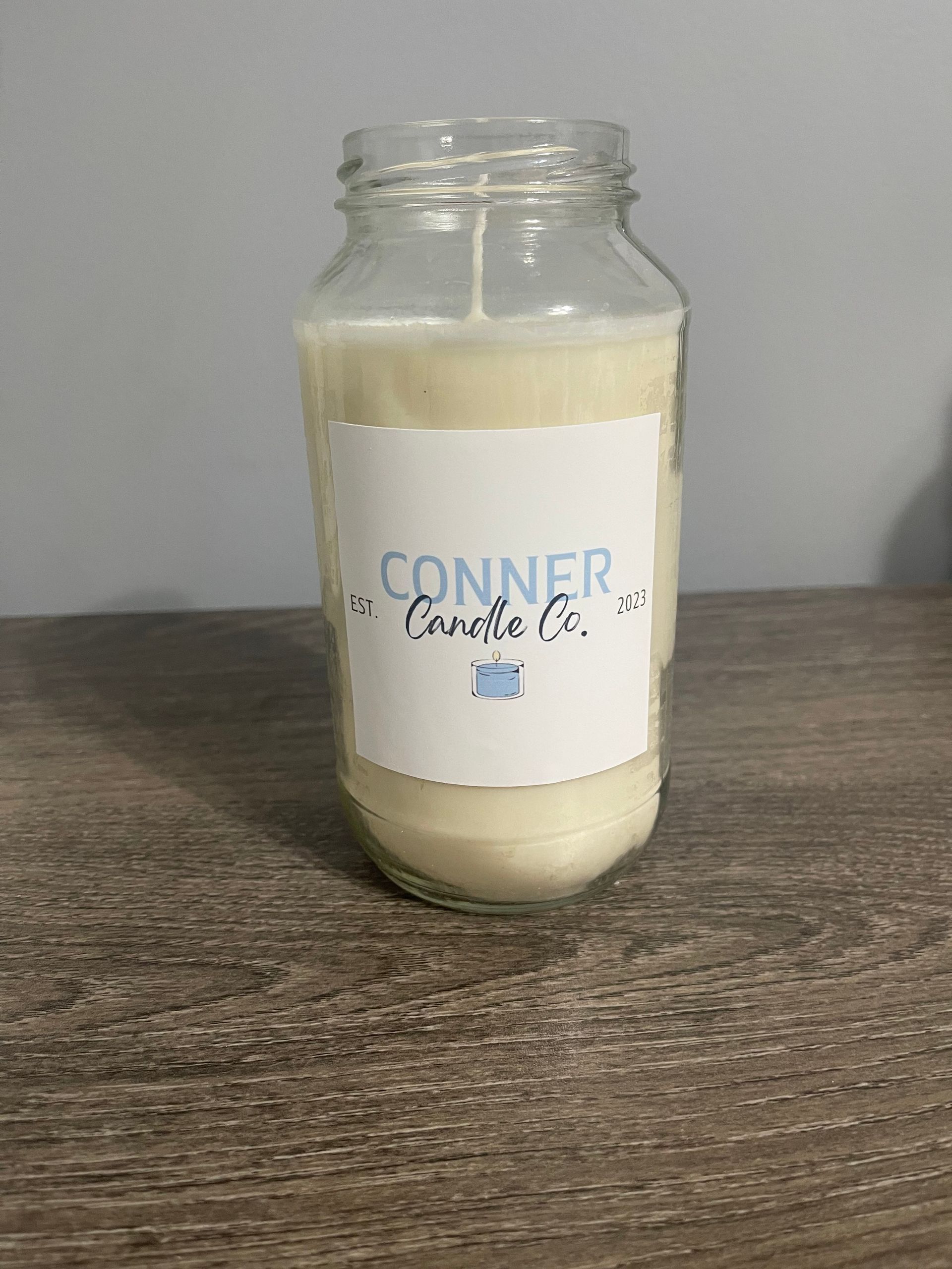 White candle in a glass jar, labeled "Conner Candle Co." on a wooden table.