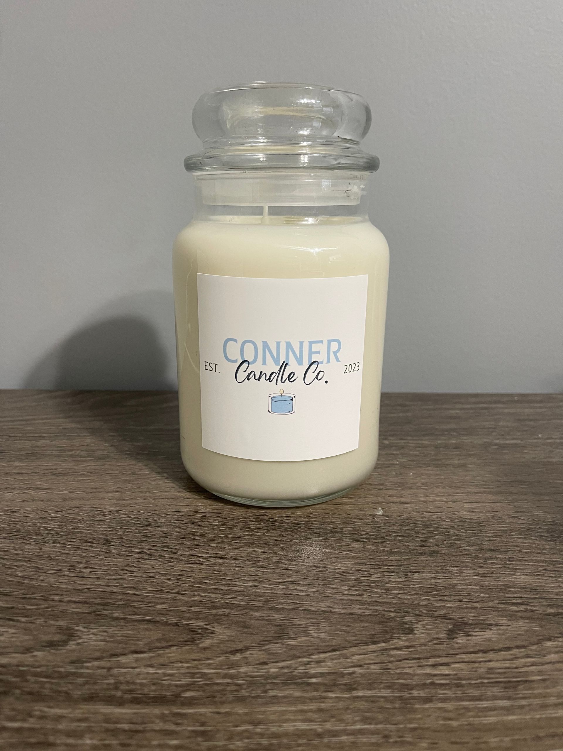 White candle in a glass jar with a lid. Label reads "Conner Candle Co." on a wood table.