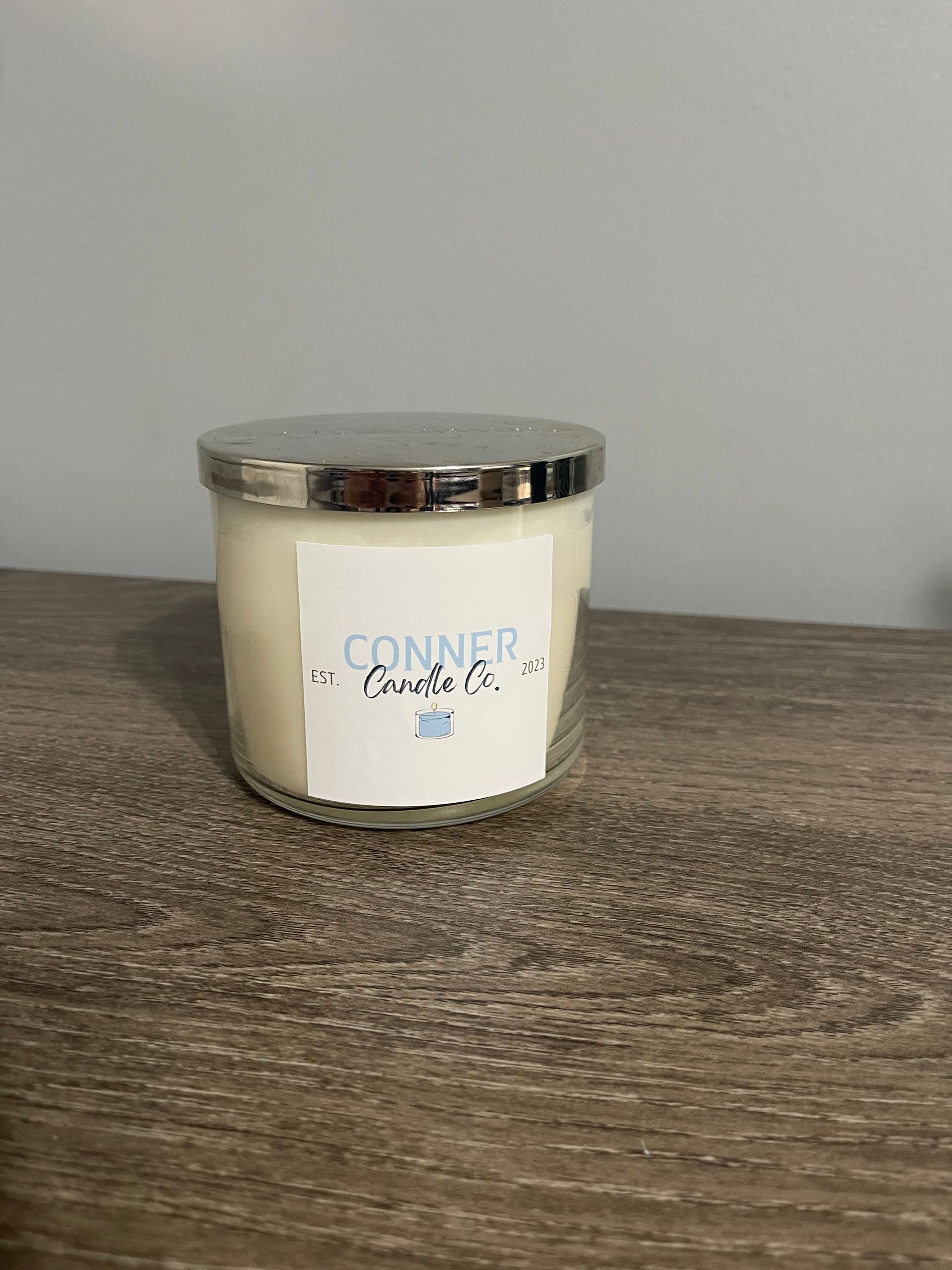 Candle in a glass jar with silver lid, labeled "CONNER Candle Co." on a wooden surface.