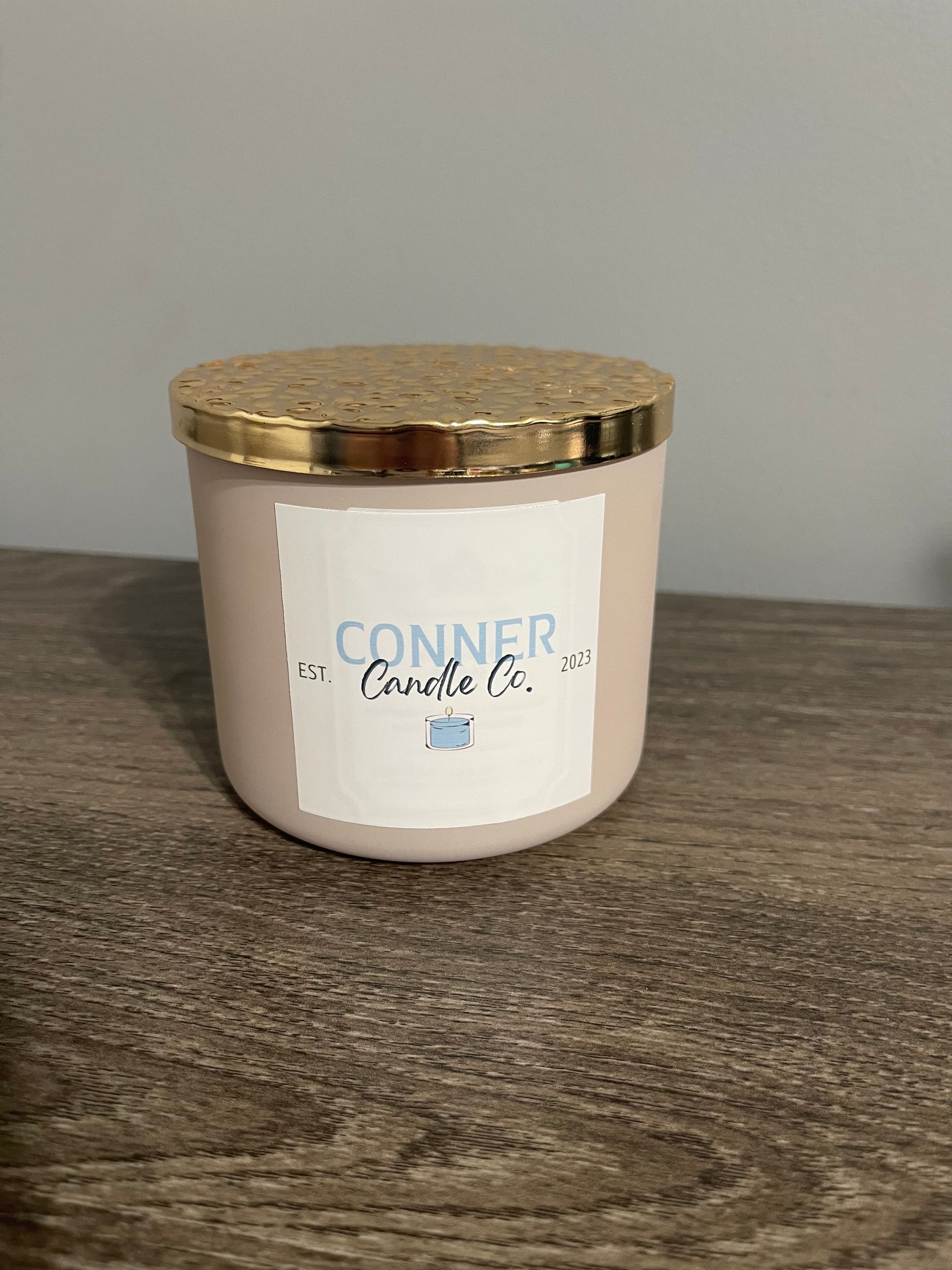 Pink candle in a jar with a gold lid, labeled "CONNER Candle Co." on a wooden surface.