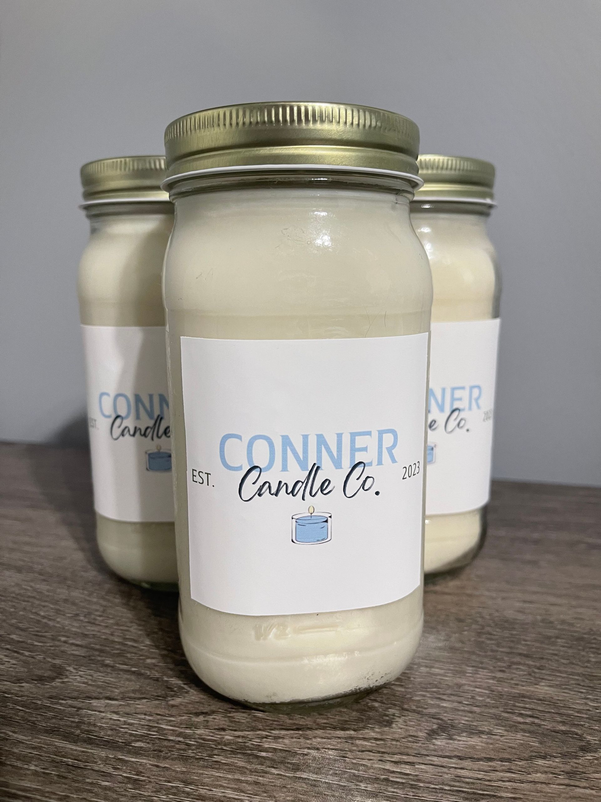 Three cream-colored jar candles with labels that read "Conner Candle Co." on a wooden surface.