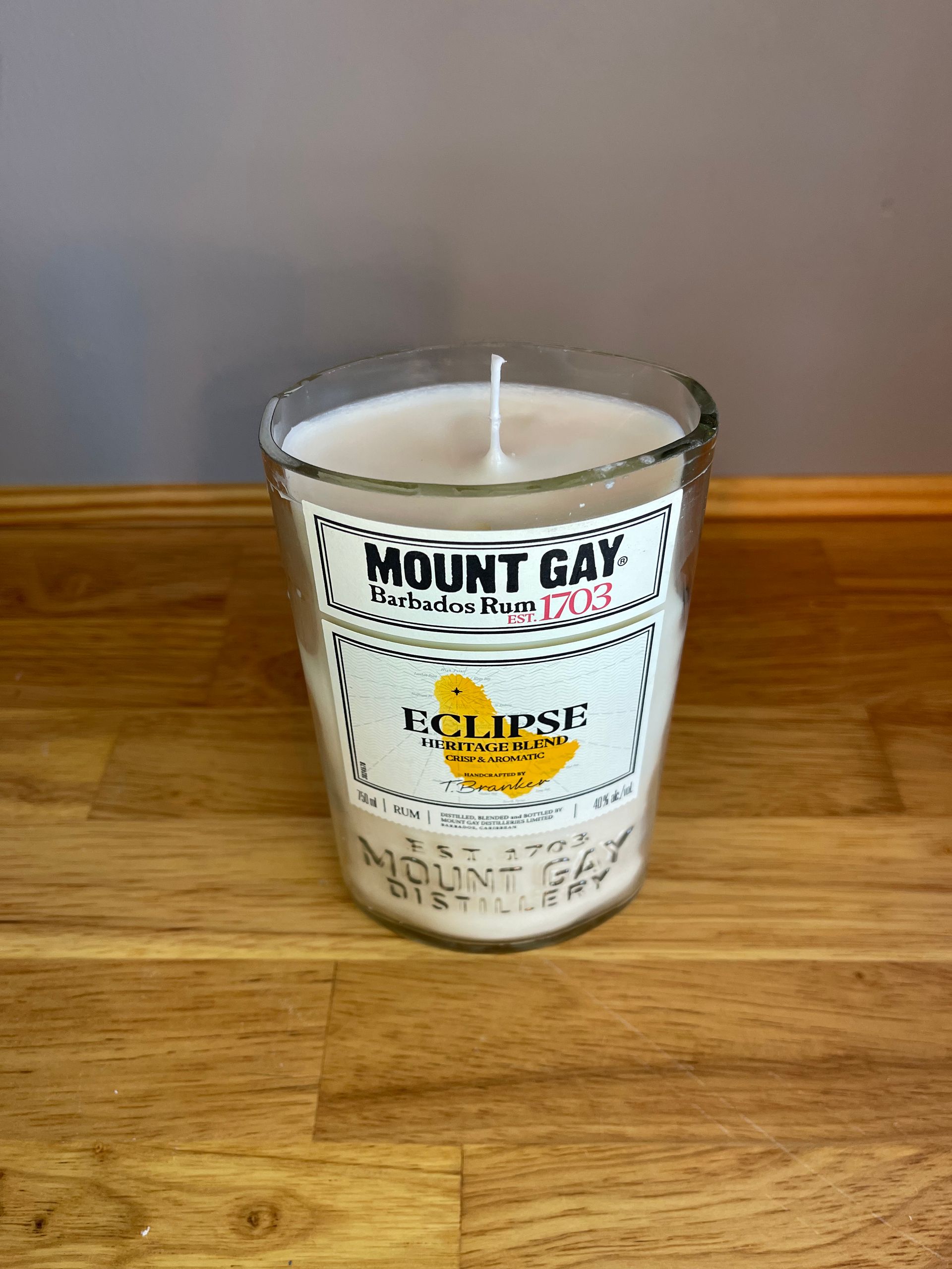 A clear glass candle on a wooden table with a Mount Gay label and yellow Eclipse wax inside