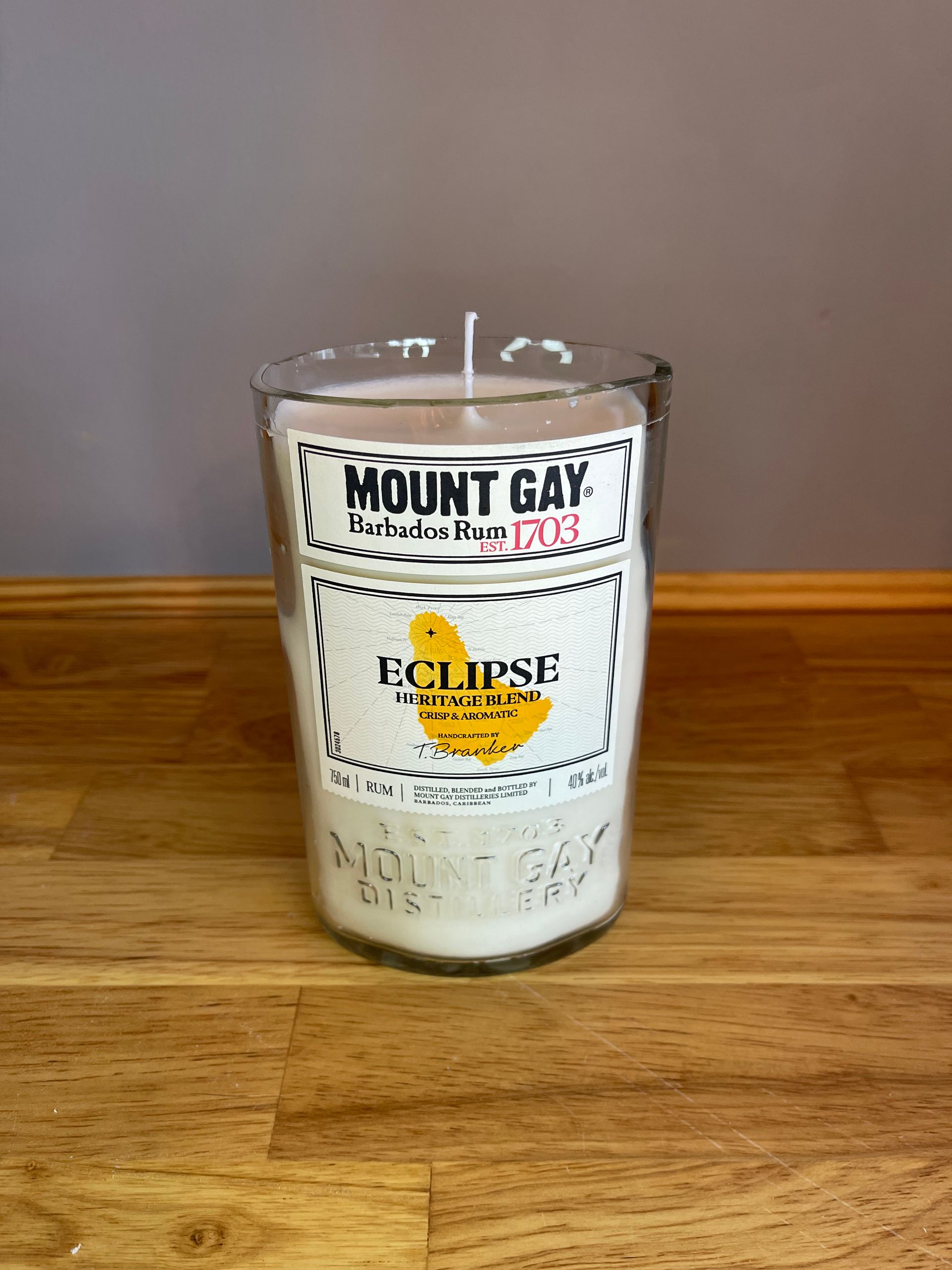 A clear glass candle on a wooden table with a Mount Gay label and yellow Eclipse wax inside