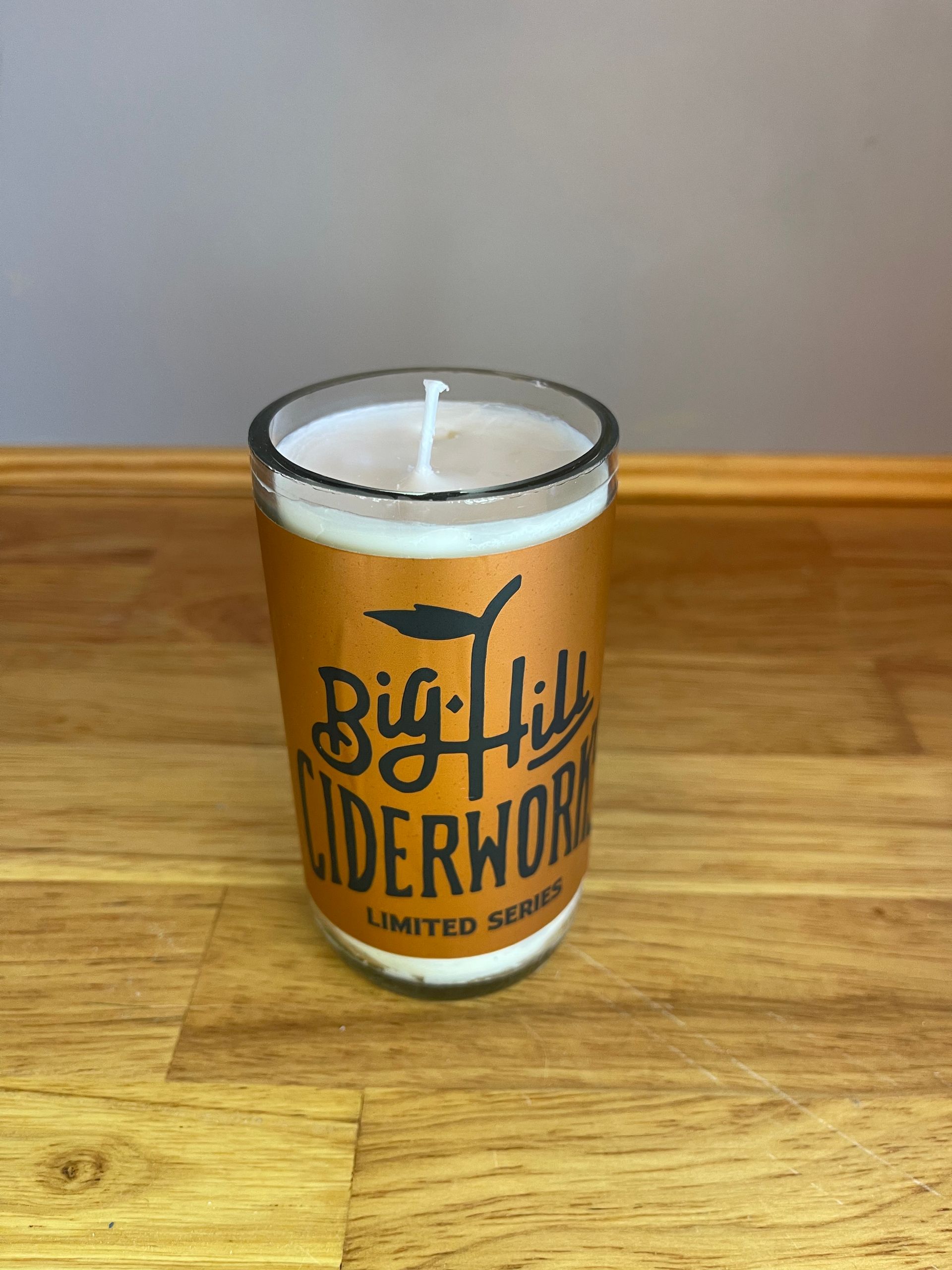 White candle in a clear glass jar with orange Big Hill Ciderworks label on a wooden surface.