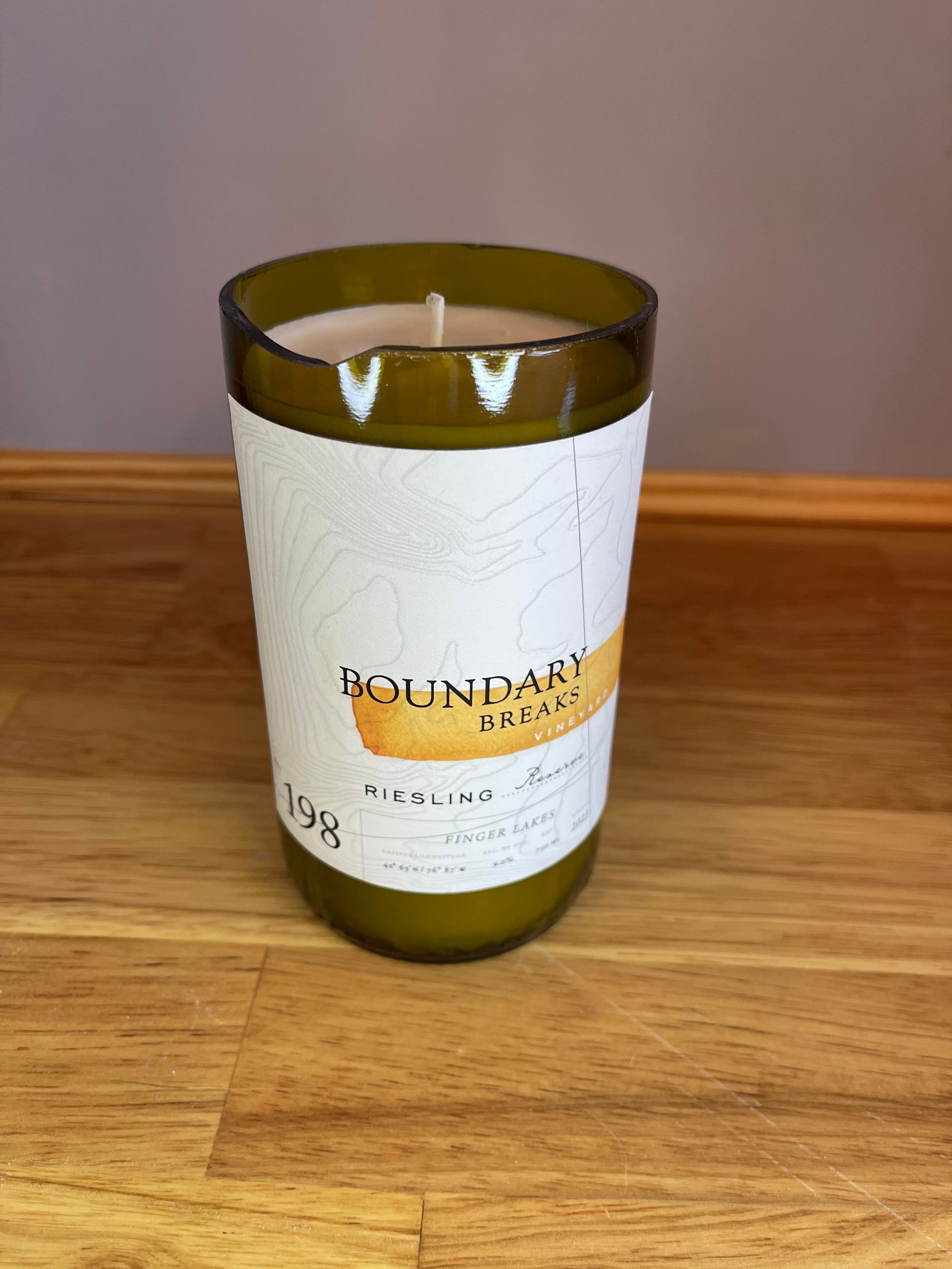 A green glass candle with a white label sits on a wooden table against a gray wall.