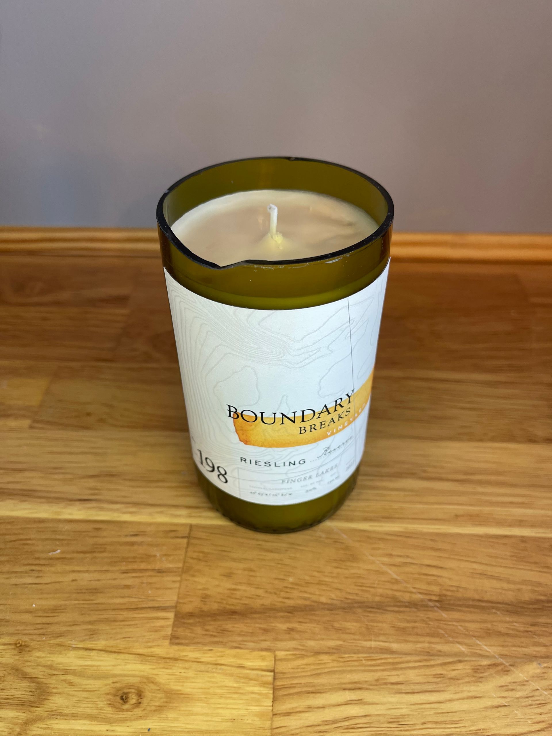 A green glass candle with a white label sits on a wooden table against a gray wall.