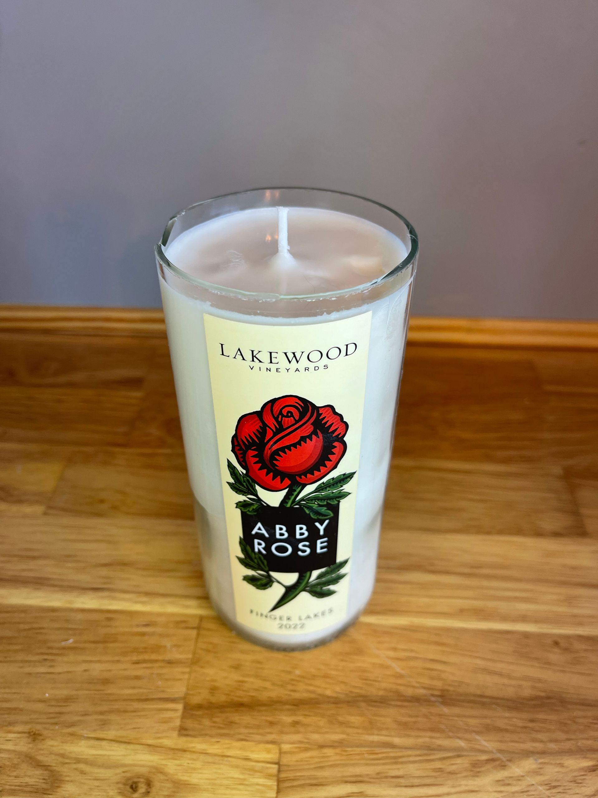 Lit candle in a clear glass with red rose and “ABBY ROSE” label on a wooden table